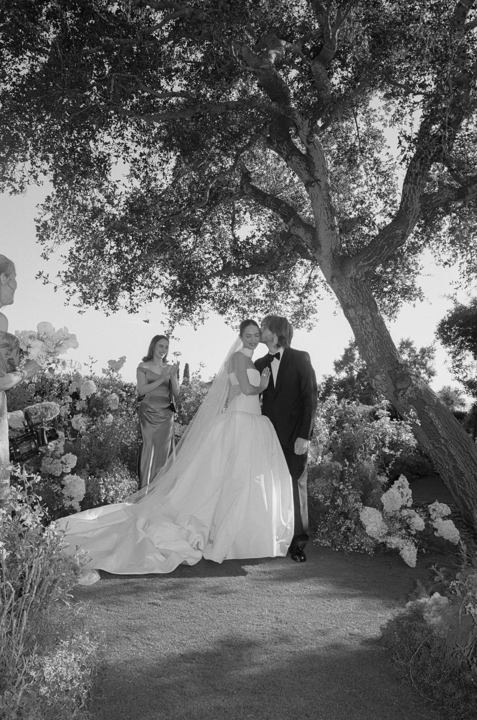 Bride and groom kiss under a large tree at an outdoor wedding, with bridesmaids and guests nearby amidst floral arrangements and the bride's long train trailing on the ground.