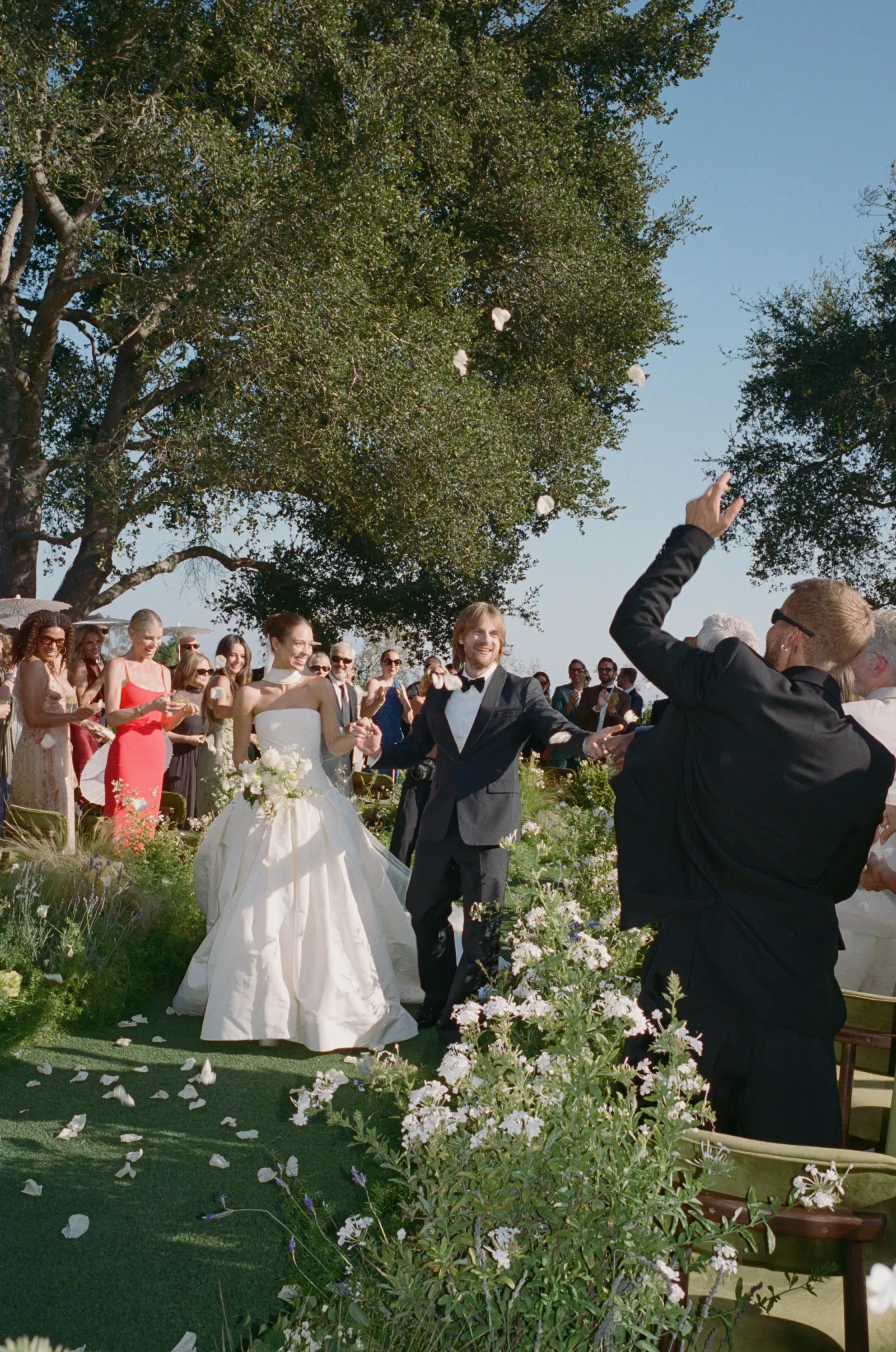 Bride in a white gown and groom in a black tuxedo stand together outdoors as petals are tossed amid cheering guests under trees.