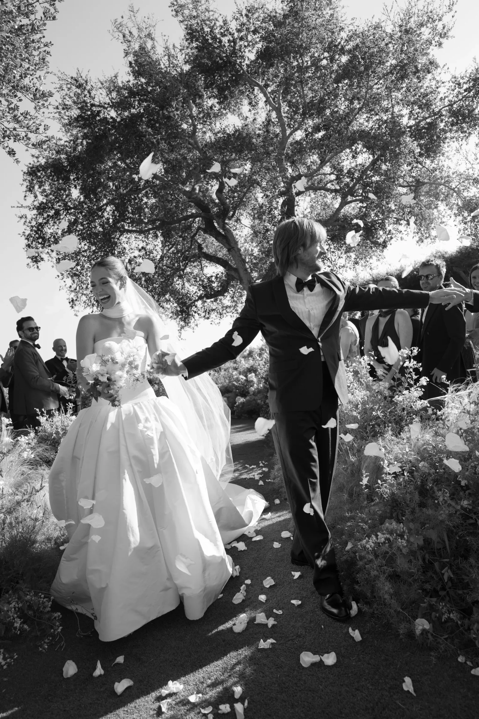 Bride and groom walk outdoors as petals scatter around them, guests cheering in the background.