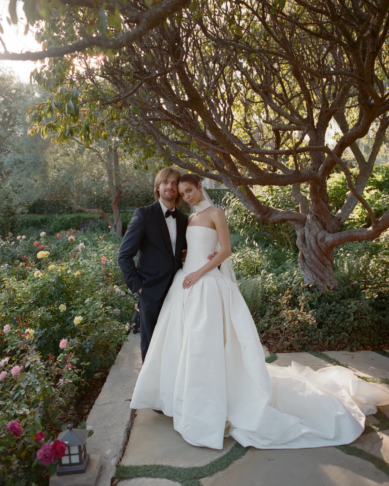 Bride in a white wedding gown and groom in a black tuxedo pose together in a garden with blooming roses and a large tree in the background.