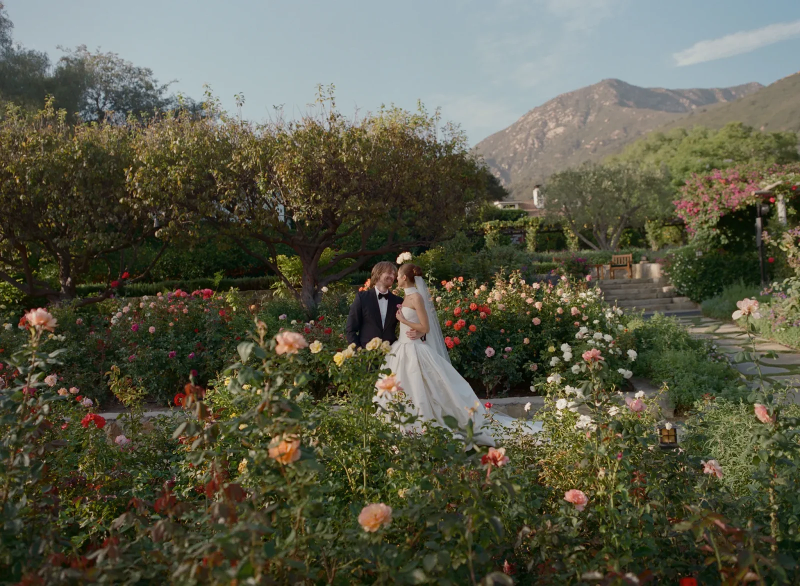 Bride and groom in wedding attire pose among blooming roses in a lush garden with mountains in the background.