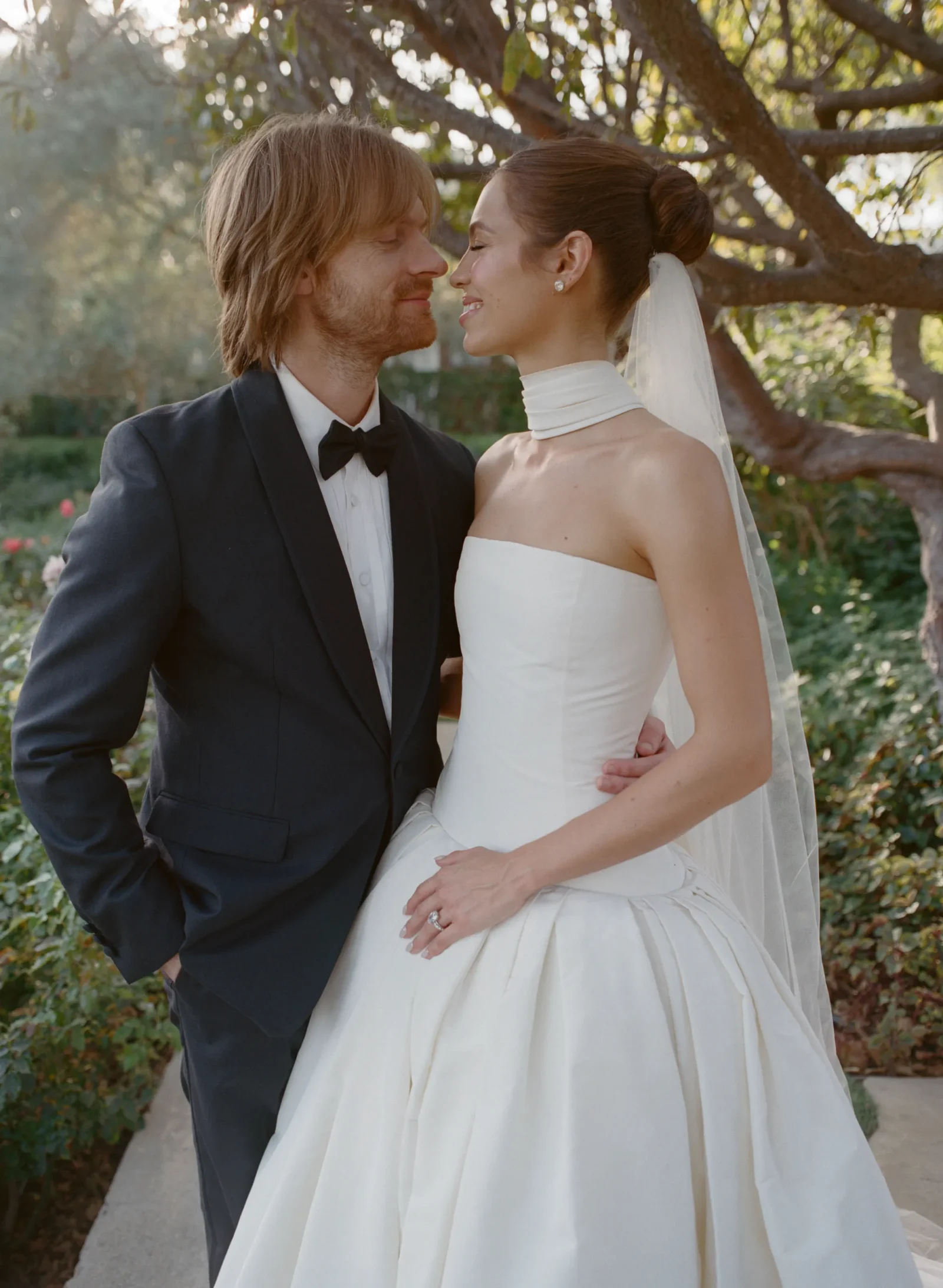 Bride and groom lean in close, smiling outdoors; she wears a white wedding dress with veil and a choker-style collar wrap? (Avoided wording)