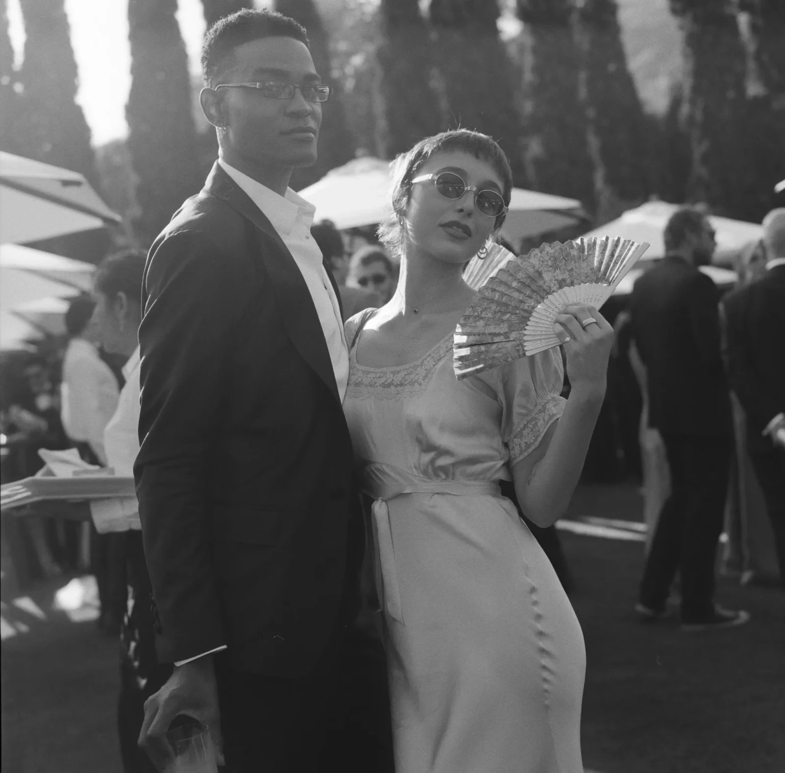 A man in a suit and a woman in a lace dress pose together at an outdoor social event; the woman holds a decorative fan.