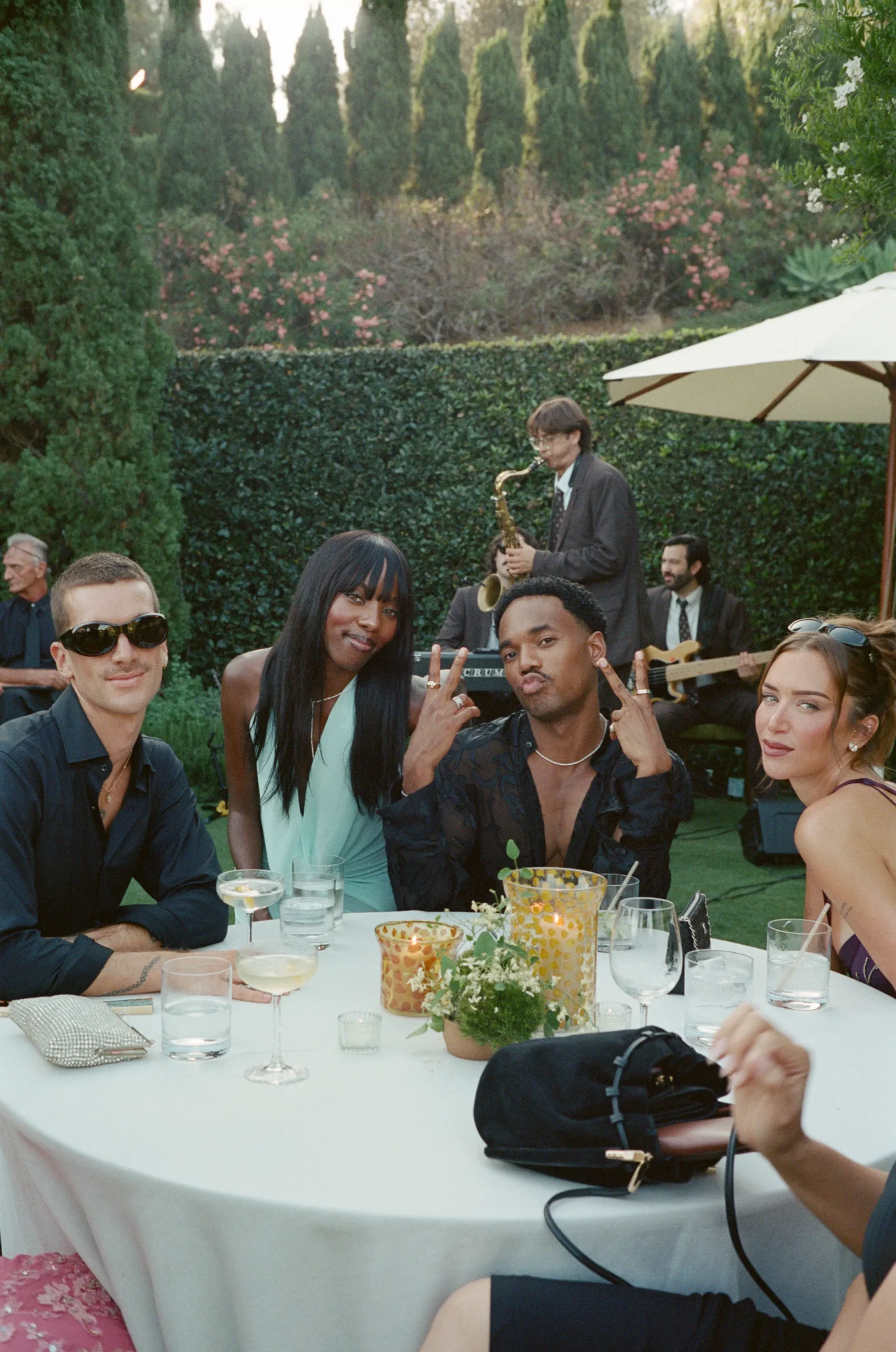 Friends at an outdoor garden party, seated at a table with drinks; a saxophonist performs in the background.