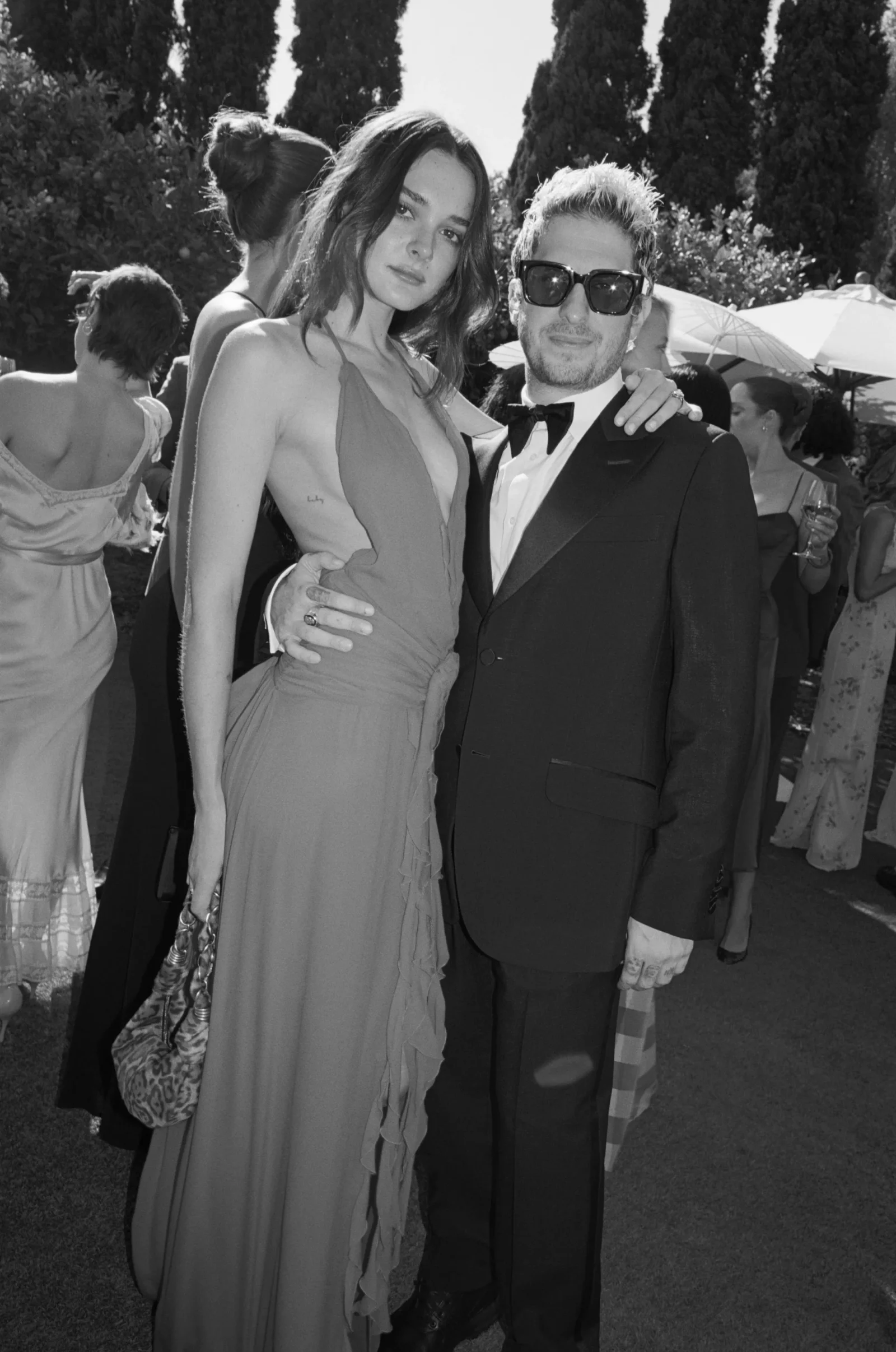 Man in a tuxedo with bow tie and sunglasses stands beside a woman in a long evening gown at an outdoor formal event, arm around her waist (black-and-white).