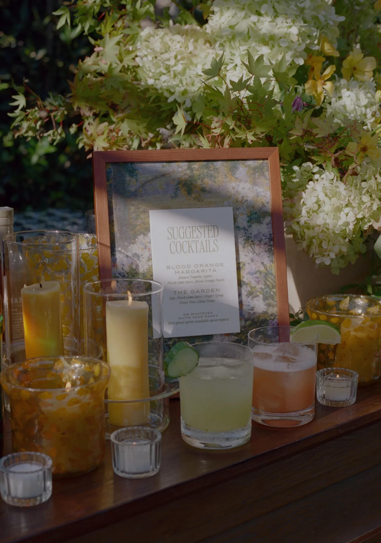 Framed menu card reading 'Suggested Cocktails' on a table with colorful drinks and a white floral backdrop.