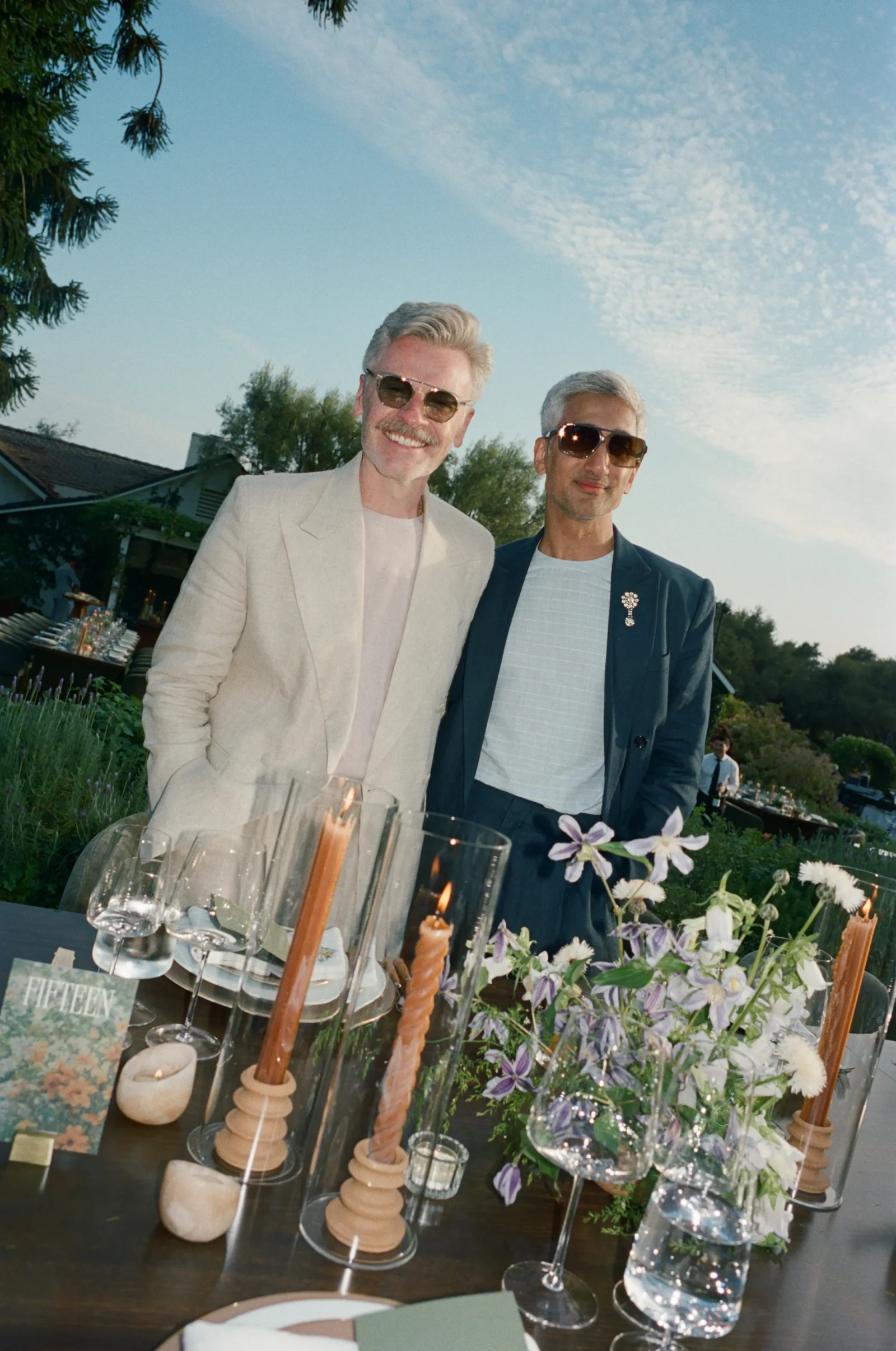 Two men in light and dark suits wearing sunglasses posing at an outdoor event table lined with candles, glassware, and floral arrangements.