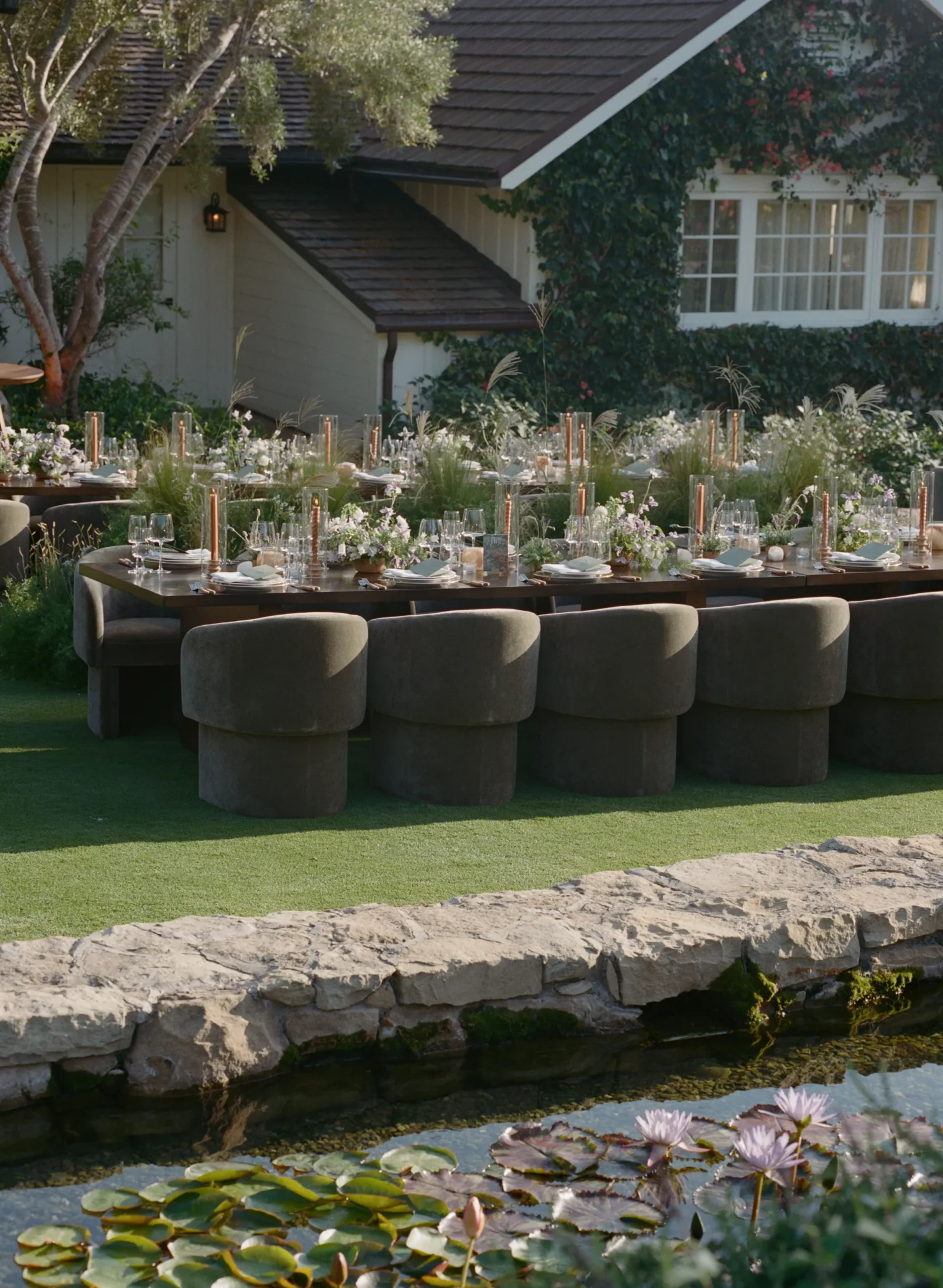 Outdoor wedding or event setup with long wooden tables, candles, glassware, and floral centerpieces beside a ivy-covered house and pond.