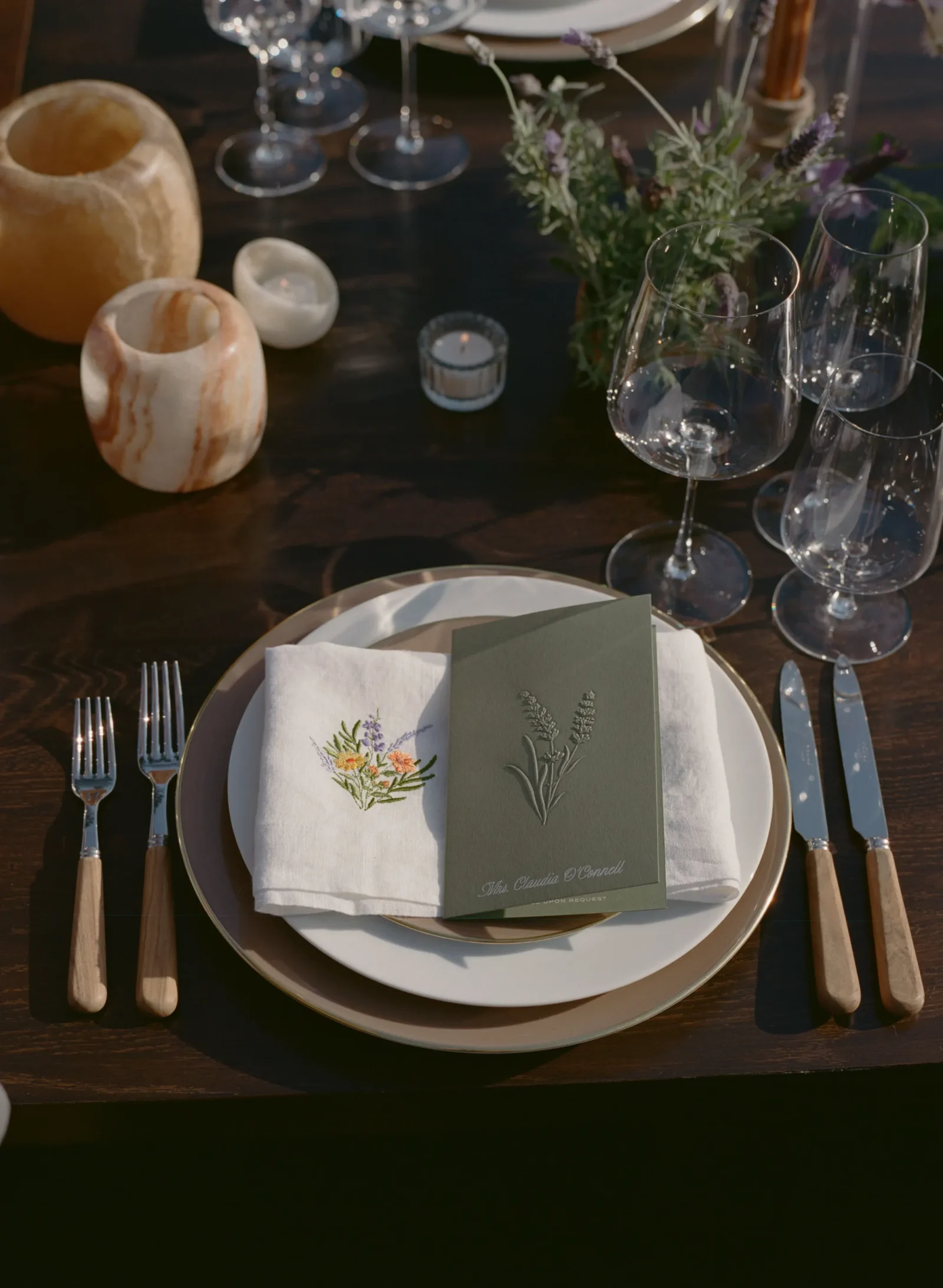 Elegant place setting with stacked plates, linen napkin featuring botanical embroidery, a green menu card, and polished cutlery on a dark wooden table with wine glasses nearby.