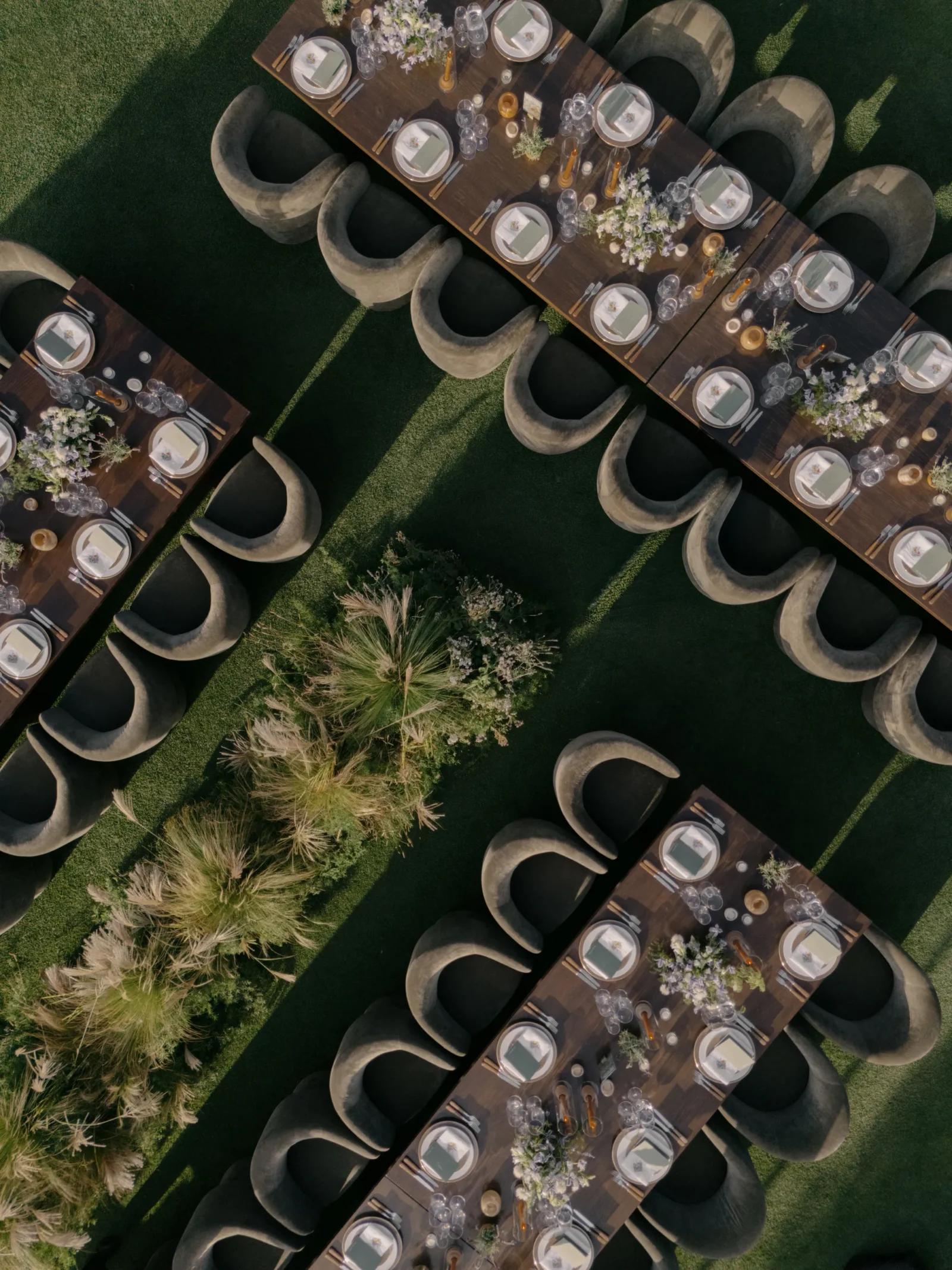 Aerial view of long wooden tables set for an outdoor event with place settings, glassware, and floral centerpieces on a grassy lawn.