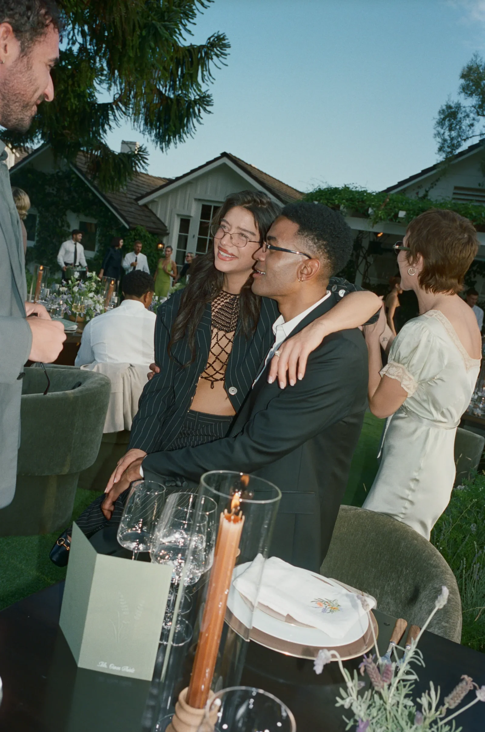 Two friends smiling and embracing at an outdoor cocktail party, with a dining table, candles, and guests in the background.