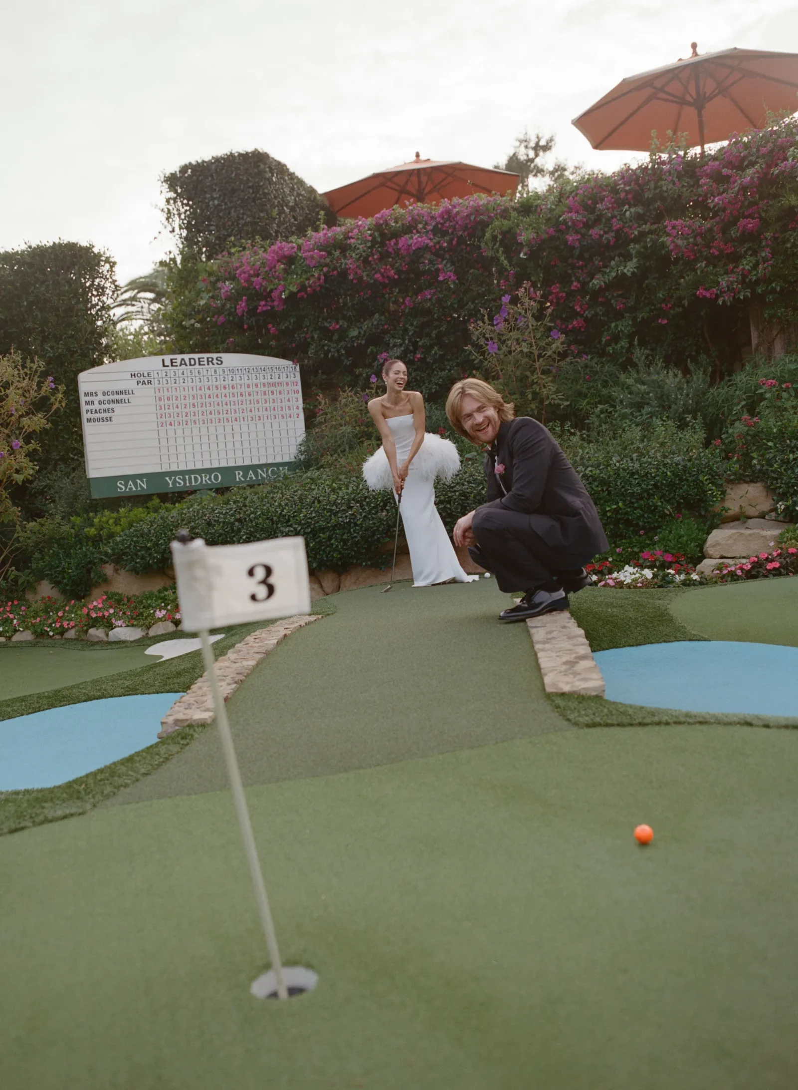 Bride in a white wedding gown laughs as a man in a dark suit crouches on a mini golf green, an orange ball nearby with a flowering hedge and scoreboard in the background.