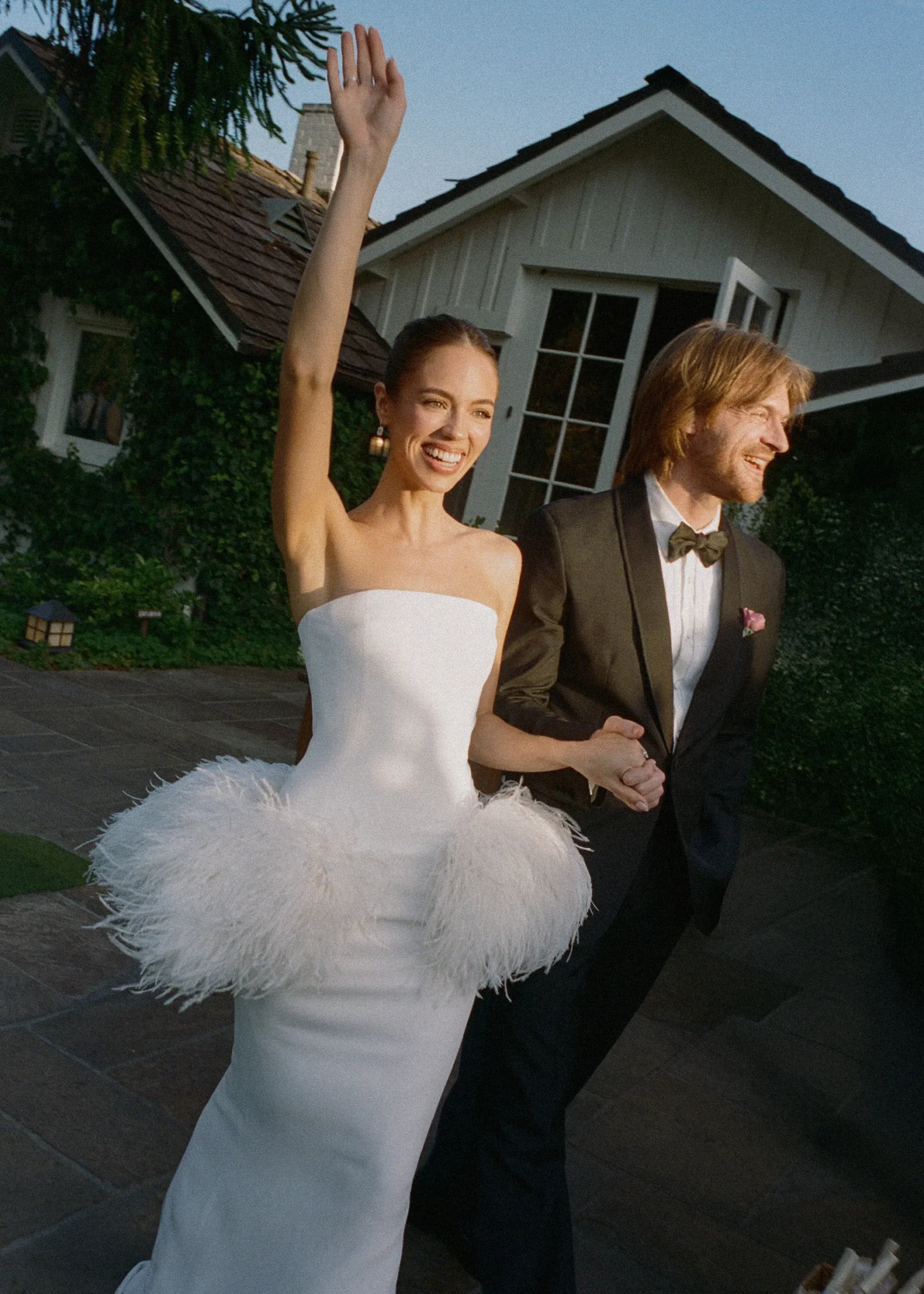 Bride in a white strapless gown waves cheerfully beside a man in a tuxedo outdoors in front of a house.