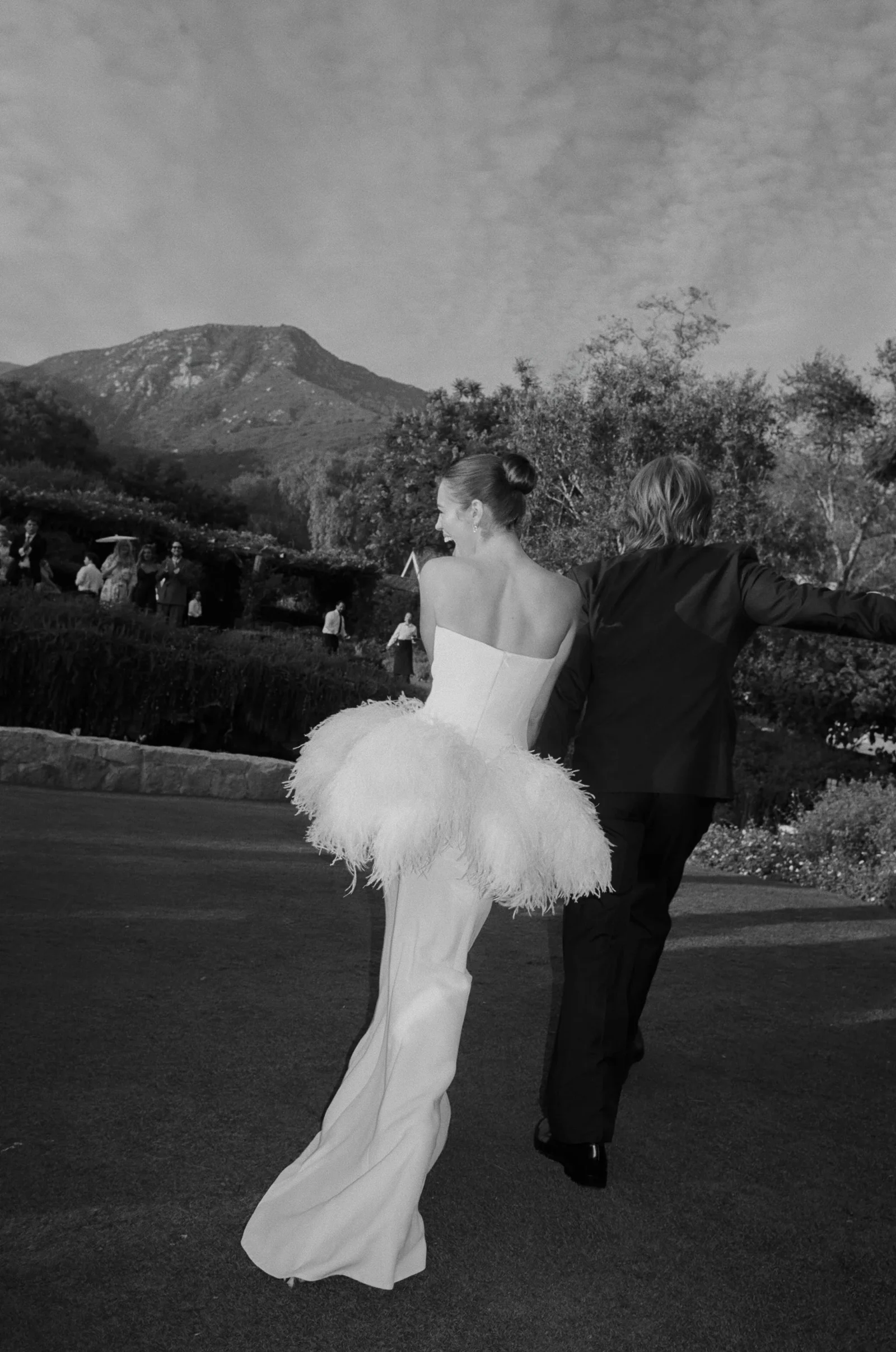 Bride in a strapless white wedding gown with a feathered skirt dancing with a man in a dark suit outdoors, mountains in the background, black-and-white photo.