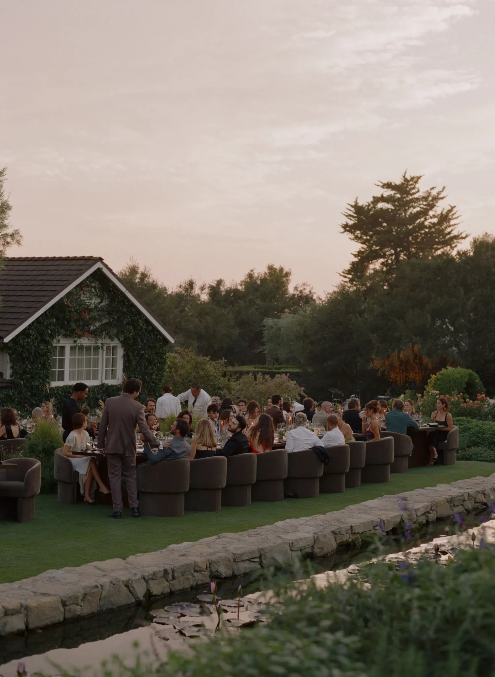 Outdoor garden dinner party at sunset with guests seated along long tables beside a ivy-covered house and stone path edge, by a pond.