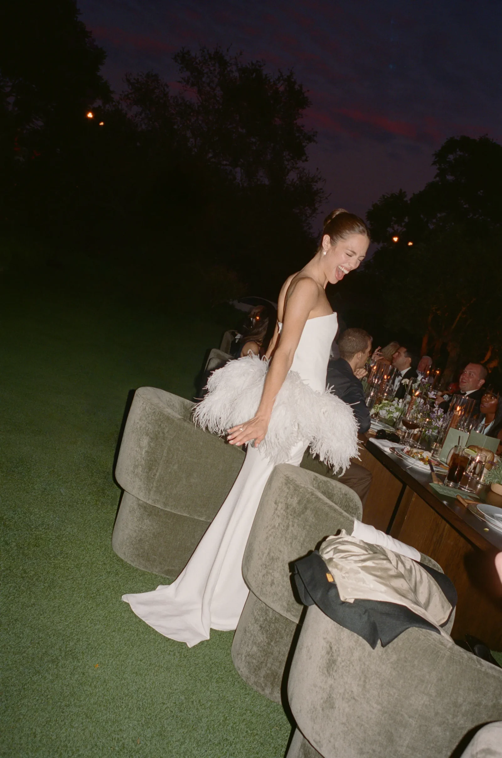 Woman in a white strapless dress with a feathered train laughing near a long outdoor dinner table at dusk garden party