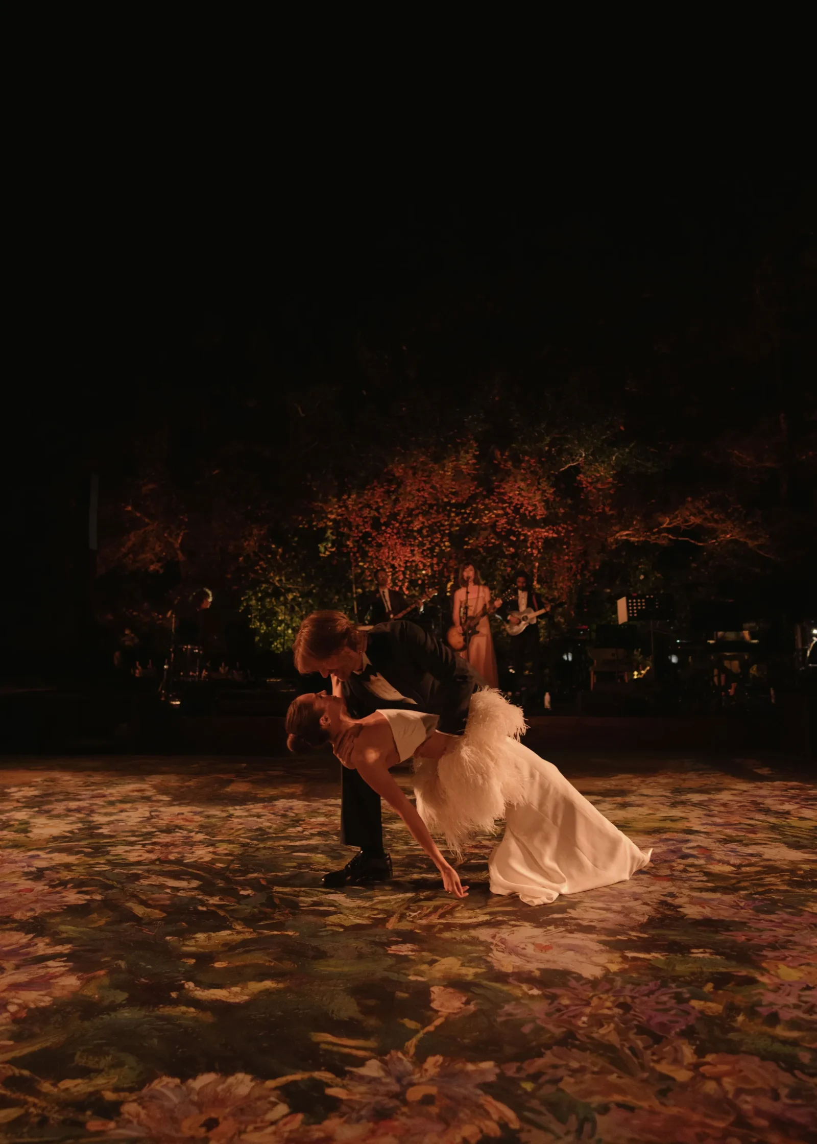 A couple dances in a dip on a dark outdoor stage, surrounded by fallen leaves and a live band in the background.