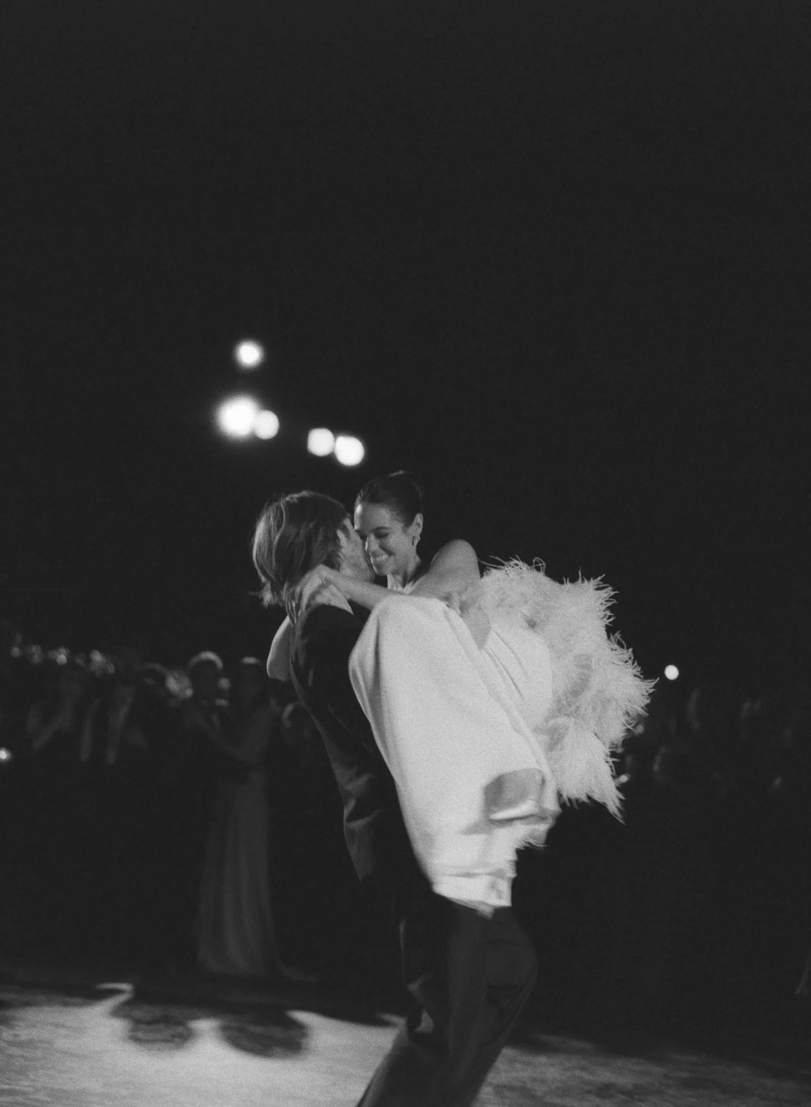 Bride in a white wedding gown being lifted by the groom while dancing at night outside.