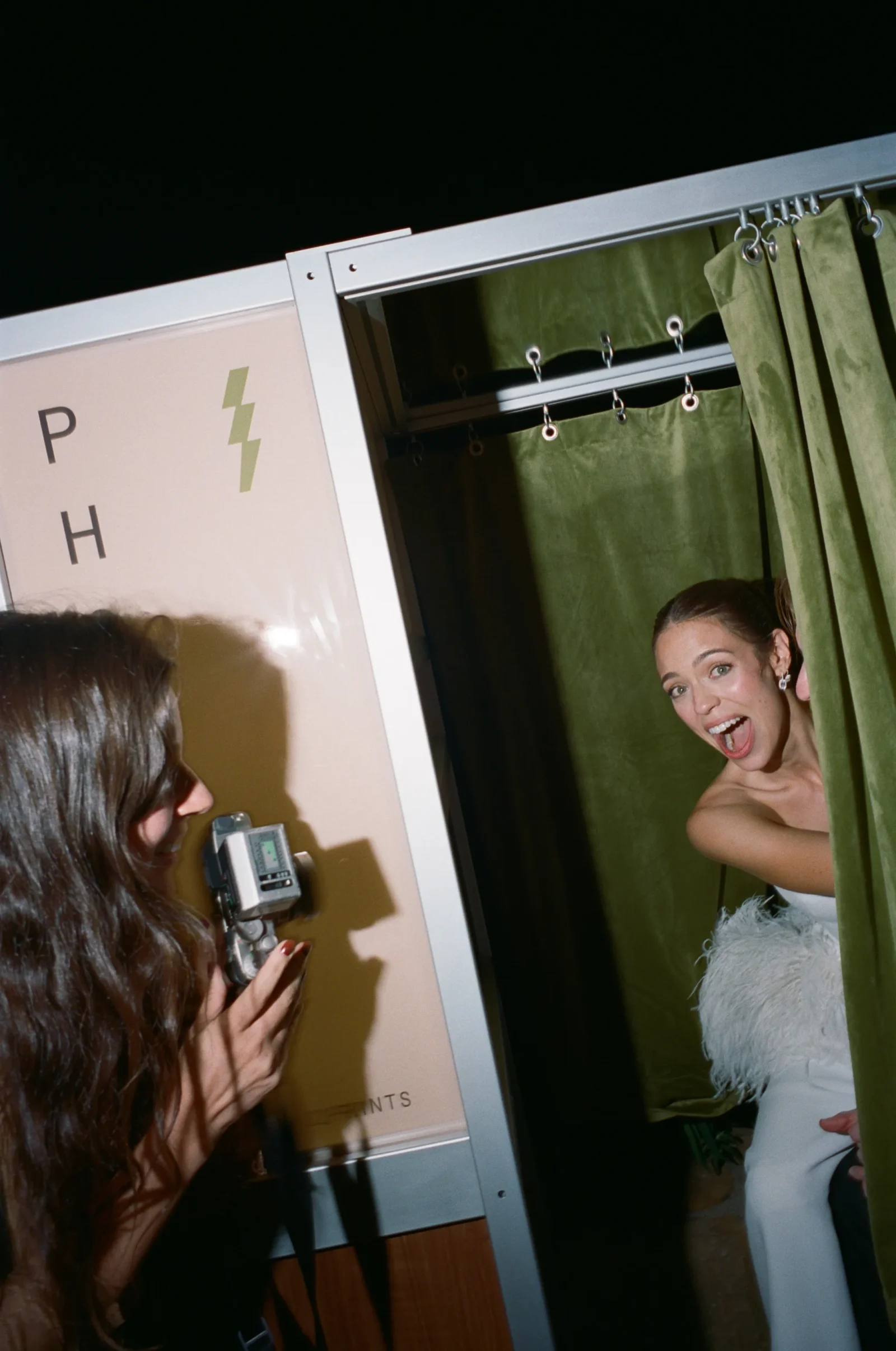 Photographer aims a flash-equipped camera at a smiling woman in a light blue dress next to green curtains in a booth setup.