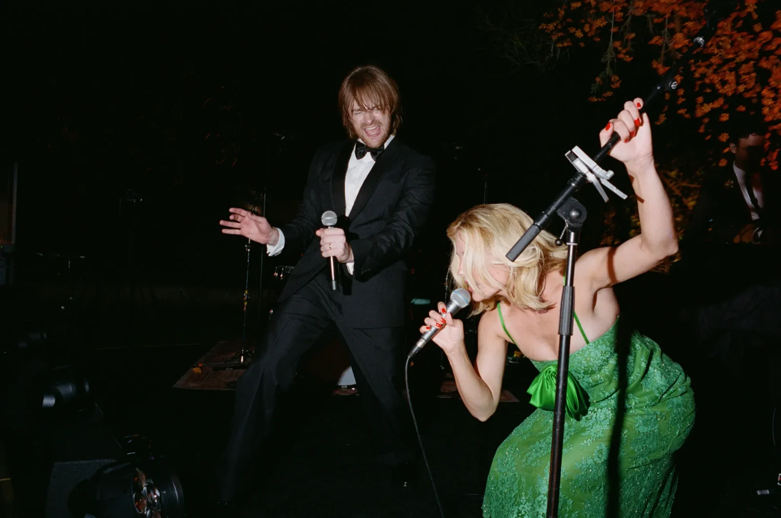 Singer in a black suit performs on stage with a microphone, next to a woman in a green dress singing into a mic.