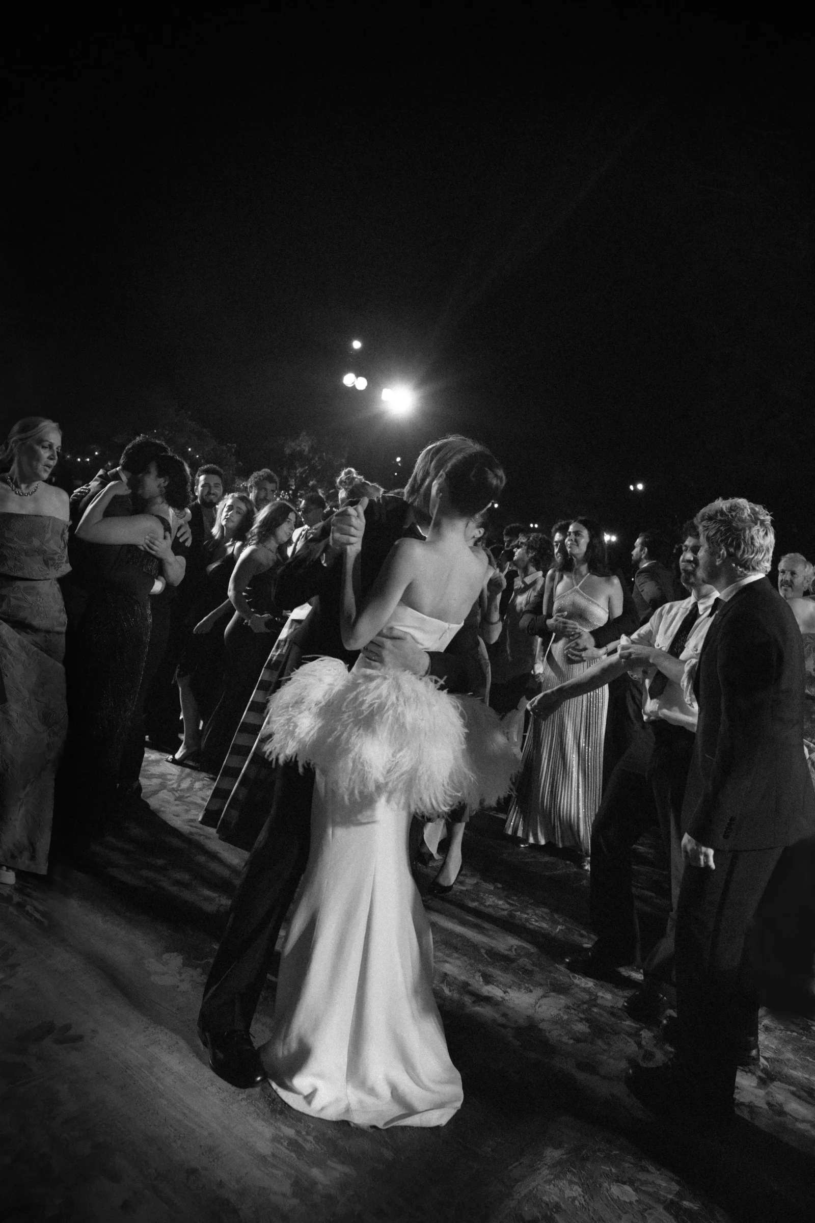 Nighttime outdoor formal dance with couples in gowns and suits; foreground shows a woman in a white gown viewed from behind.