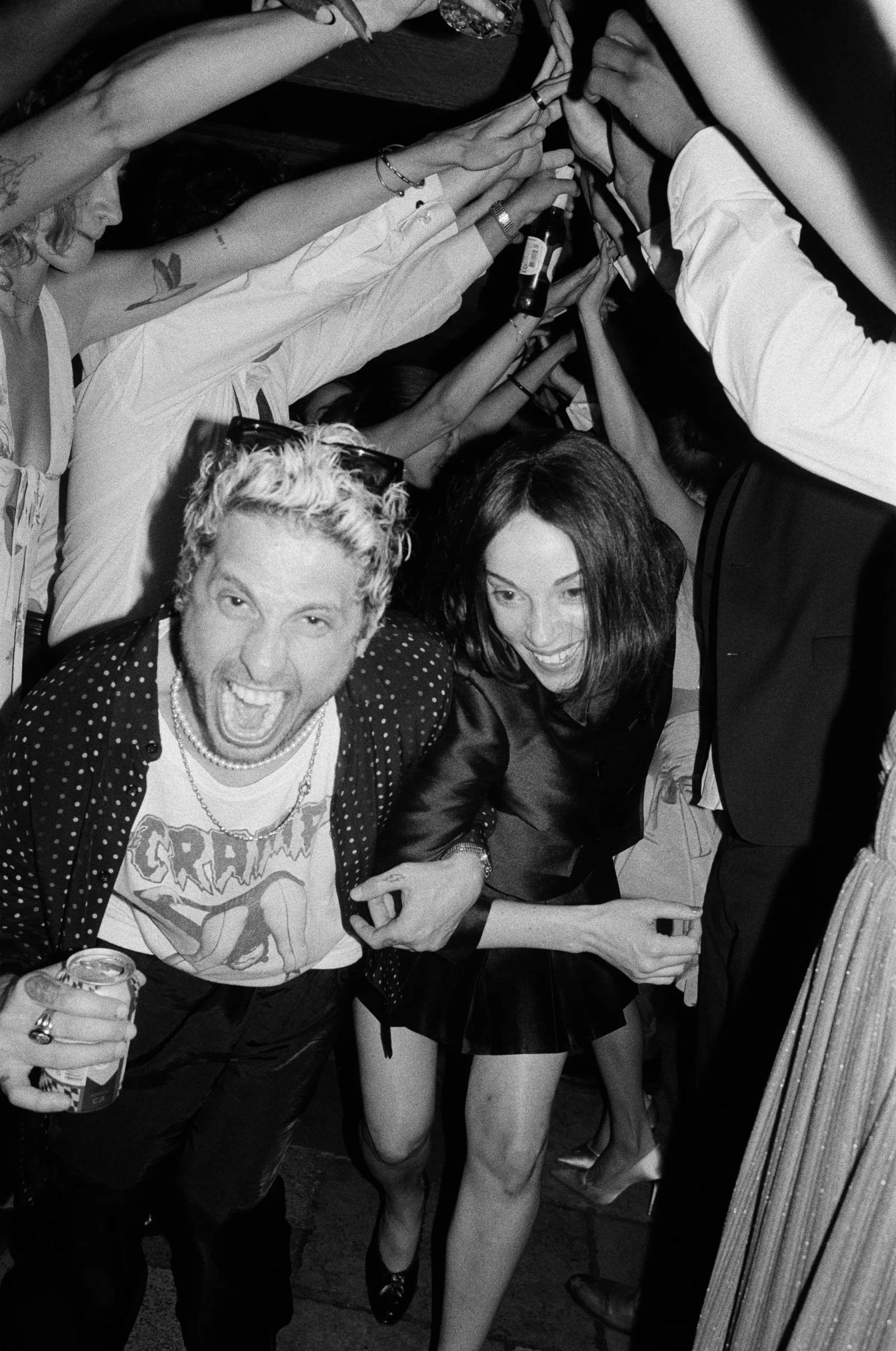 A crowded party scene with everyone raising drinks and hands toward the center, foreground showing a man with bleached hair shouting and a smiling woman beside him holding a drink.