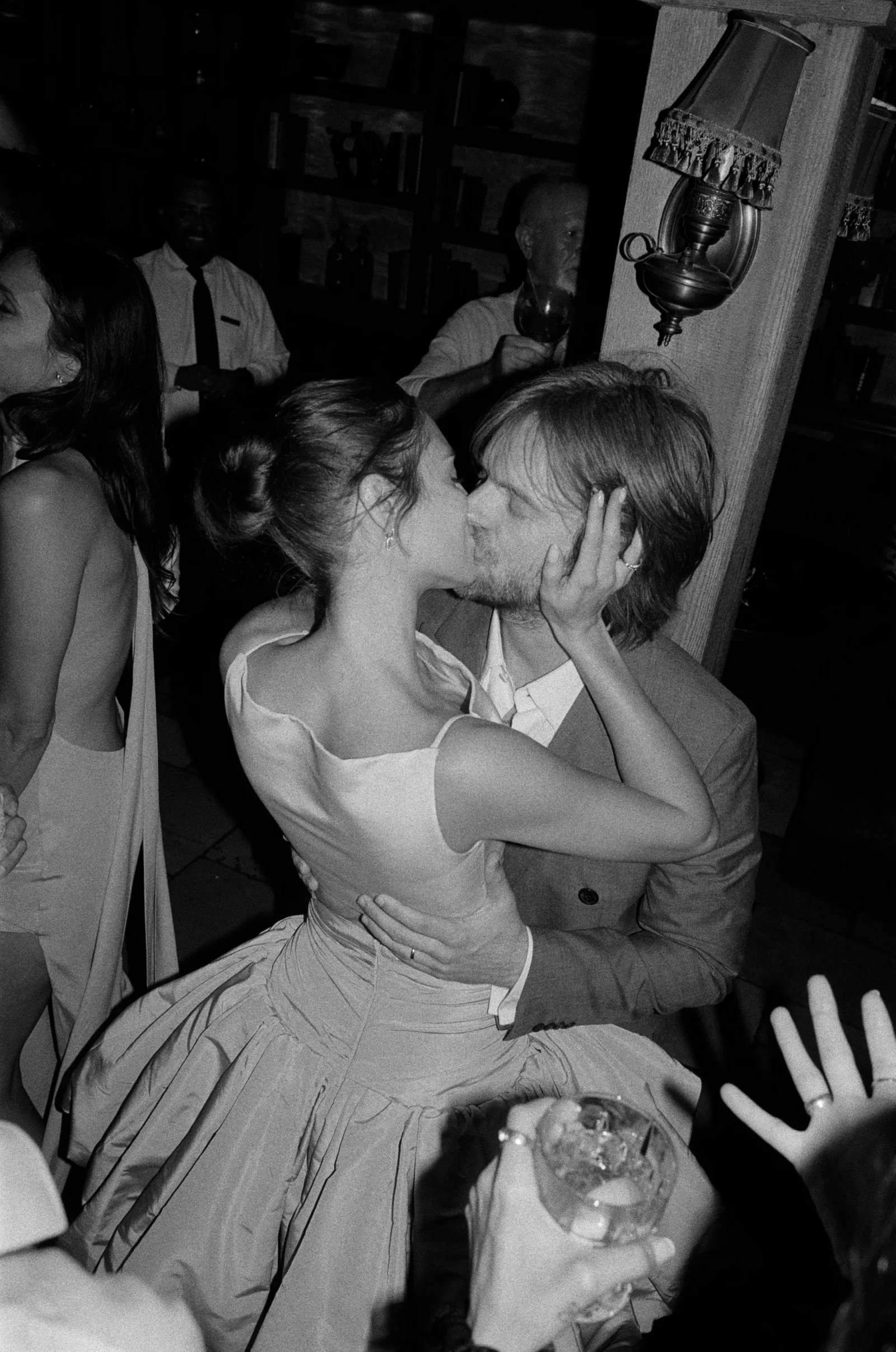 Couple kissing at a party, the woman in a gown and the man in a suit, embraced amid guests in a dimly lit room.