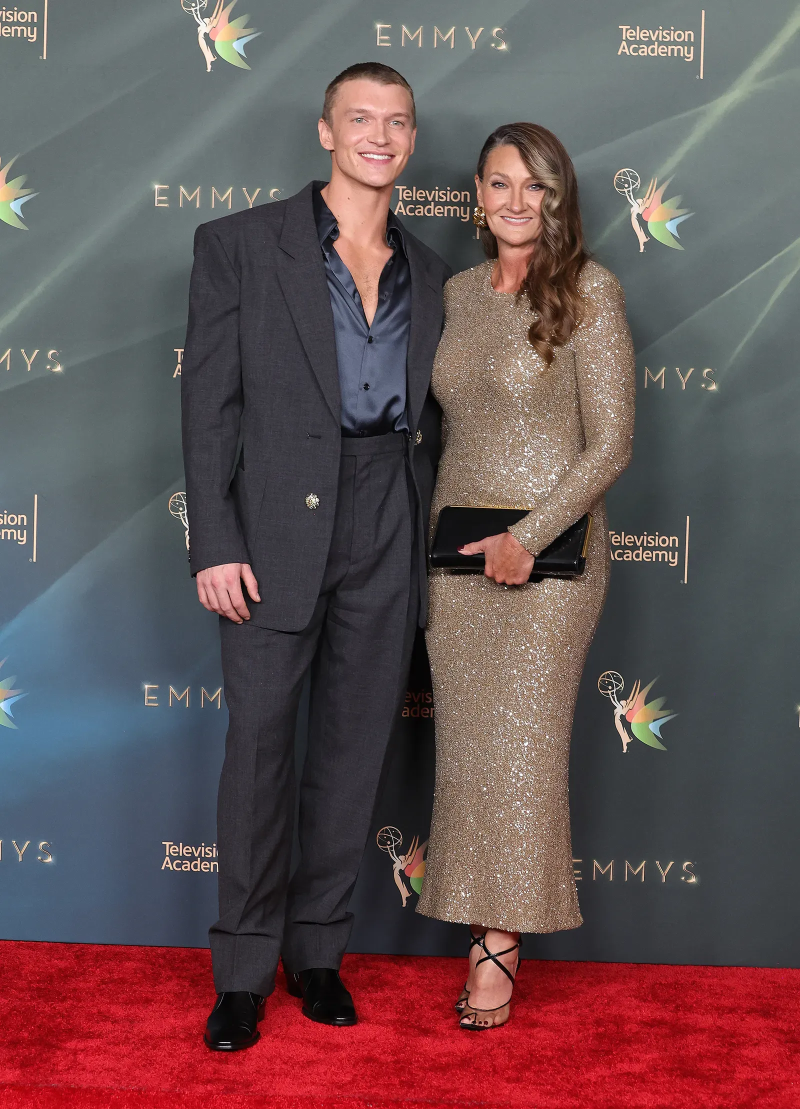 Man in a dark suit and open navy shirt posing with a smiling woman in a gold sequined dress on the Emmy Awards red carpet, with a teal backdrop behind them.