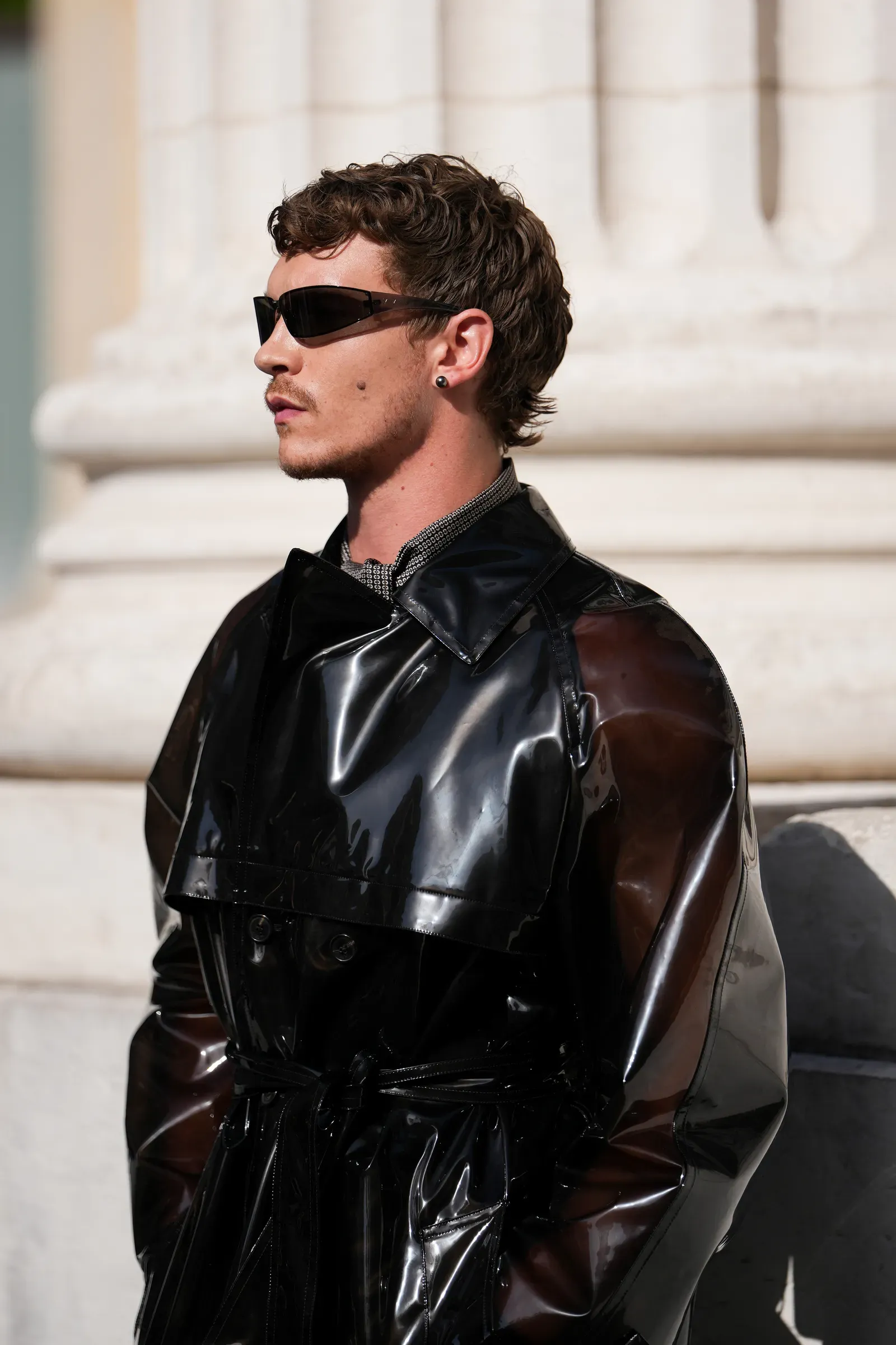 Man in a glossy black trench coat and sunglasses, standing outdoors with a stone building in the background.