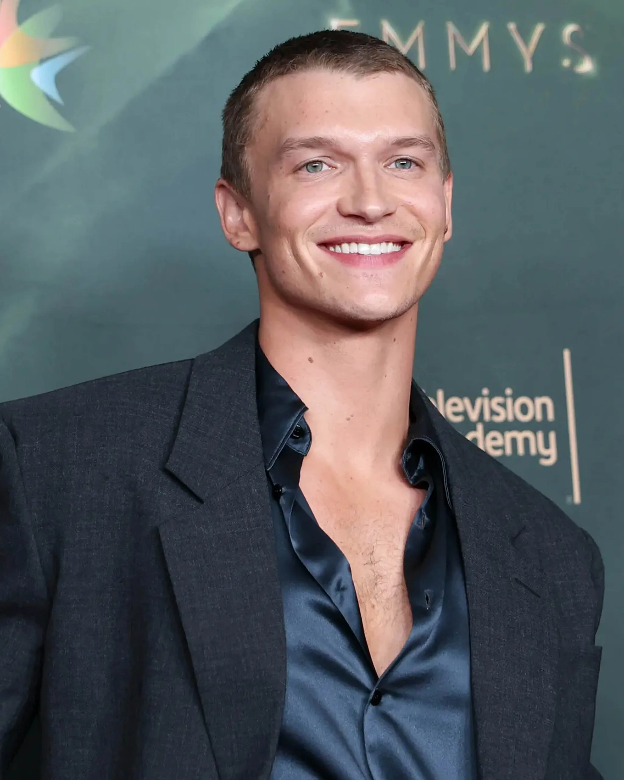 Smiling man in a dark blazer at a formal event, backdrop with logos visible behind him.