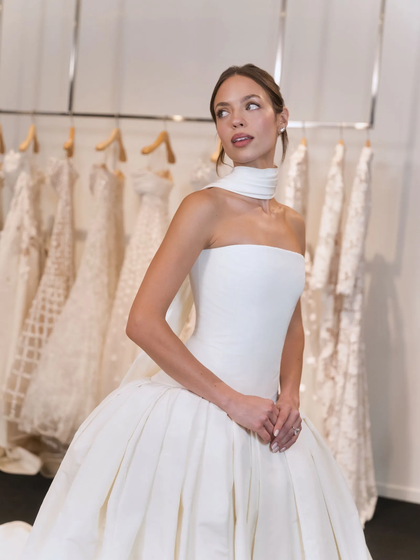 Bride-to-be in a white strapless wedding gown, standing in a bridal shop with dresses on racks in the background.
