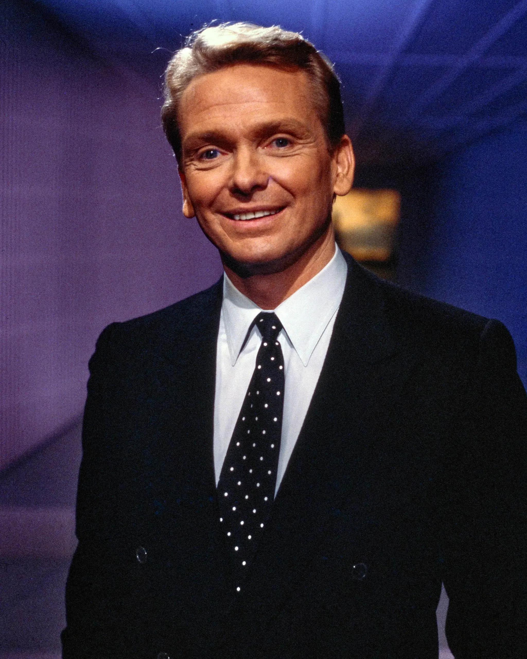 Smiling man in a dark suit and polka-dot tie, standing against a purple-blue stage background.”,