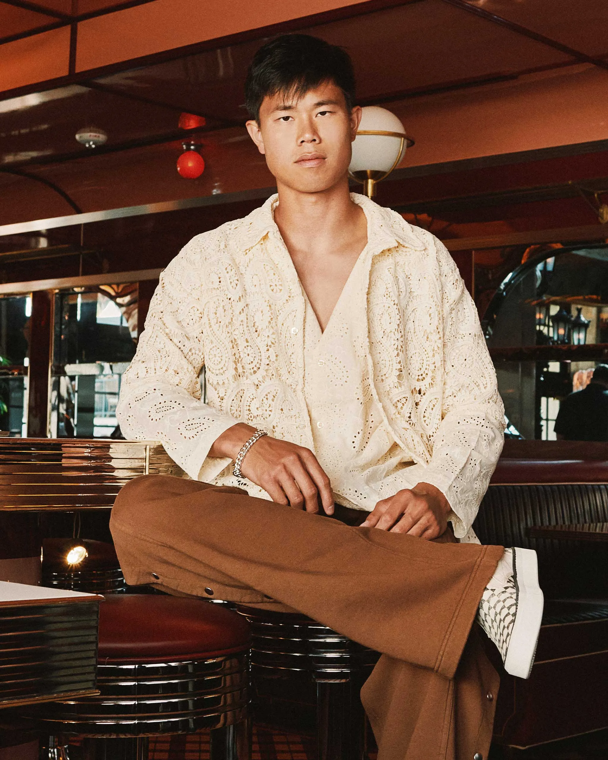 Man wearing a cream lace shirt and tan pants sits cross-legged at a bar, looking at the camera.