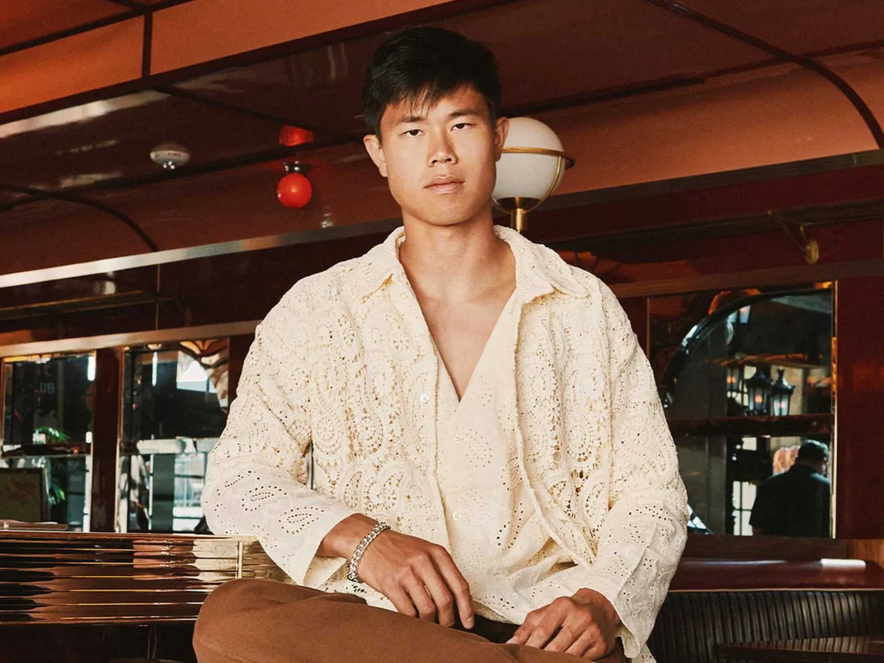 Man wearing a cream lace shirt stands in a warm, retro-looking bar, facing the camera.