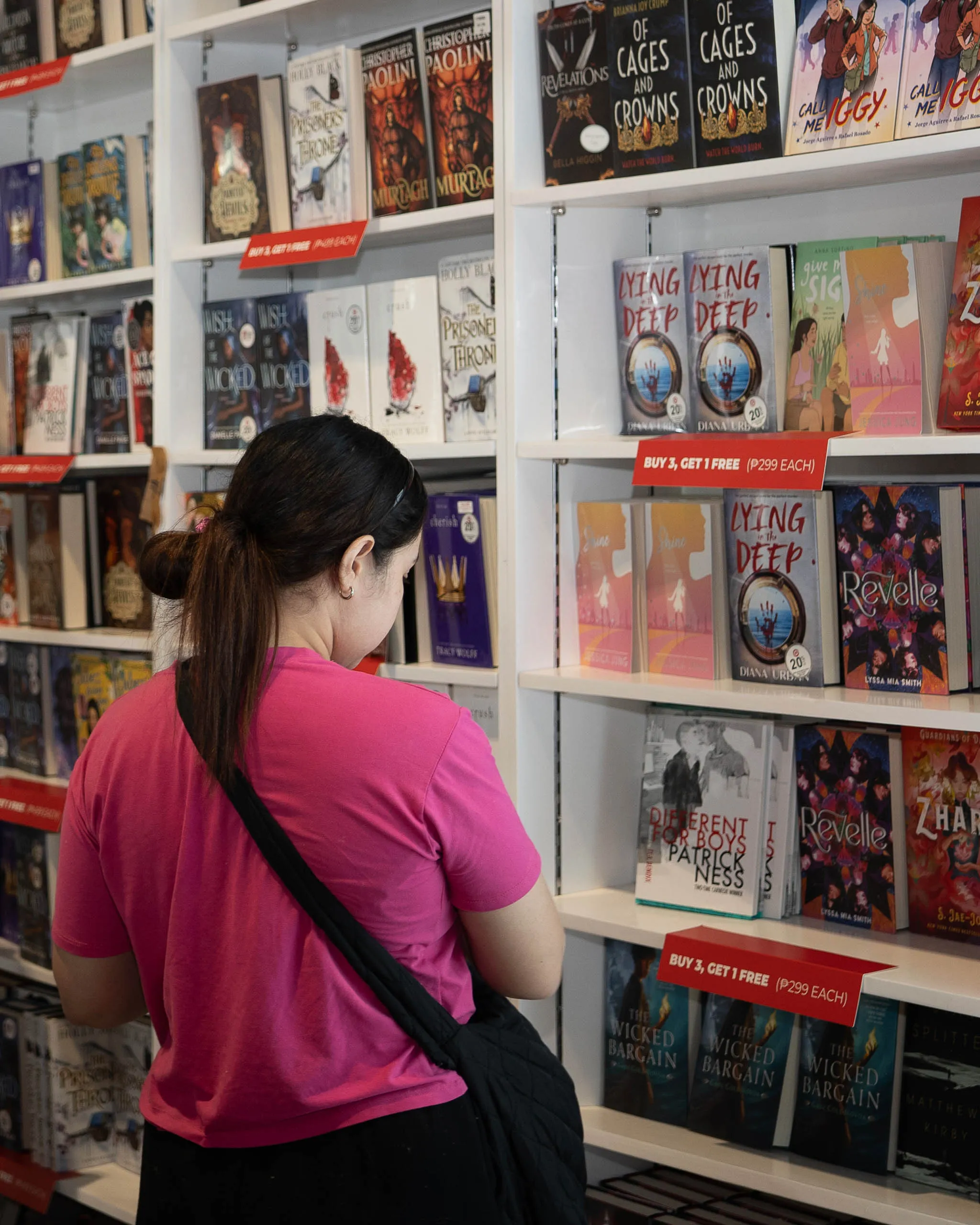 Woman in a pink shirt browsing shelves of books at a bookstore, facing away from the camera.