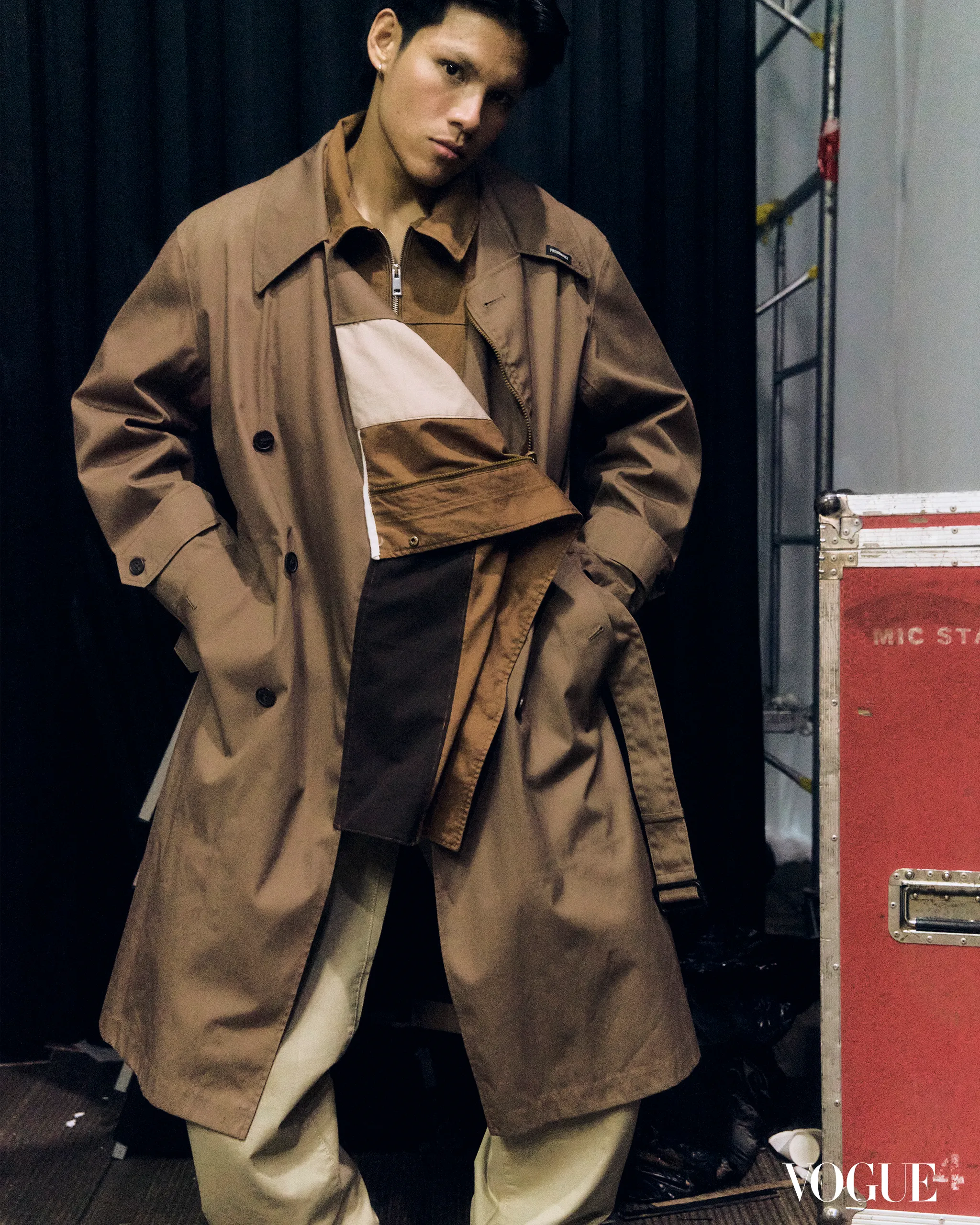 Male model in a tan trench coat posing backstage beside a red equipment case, black curtain behind.