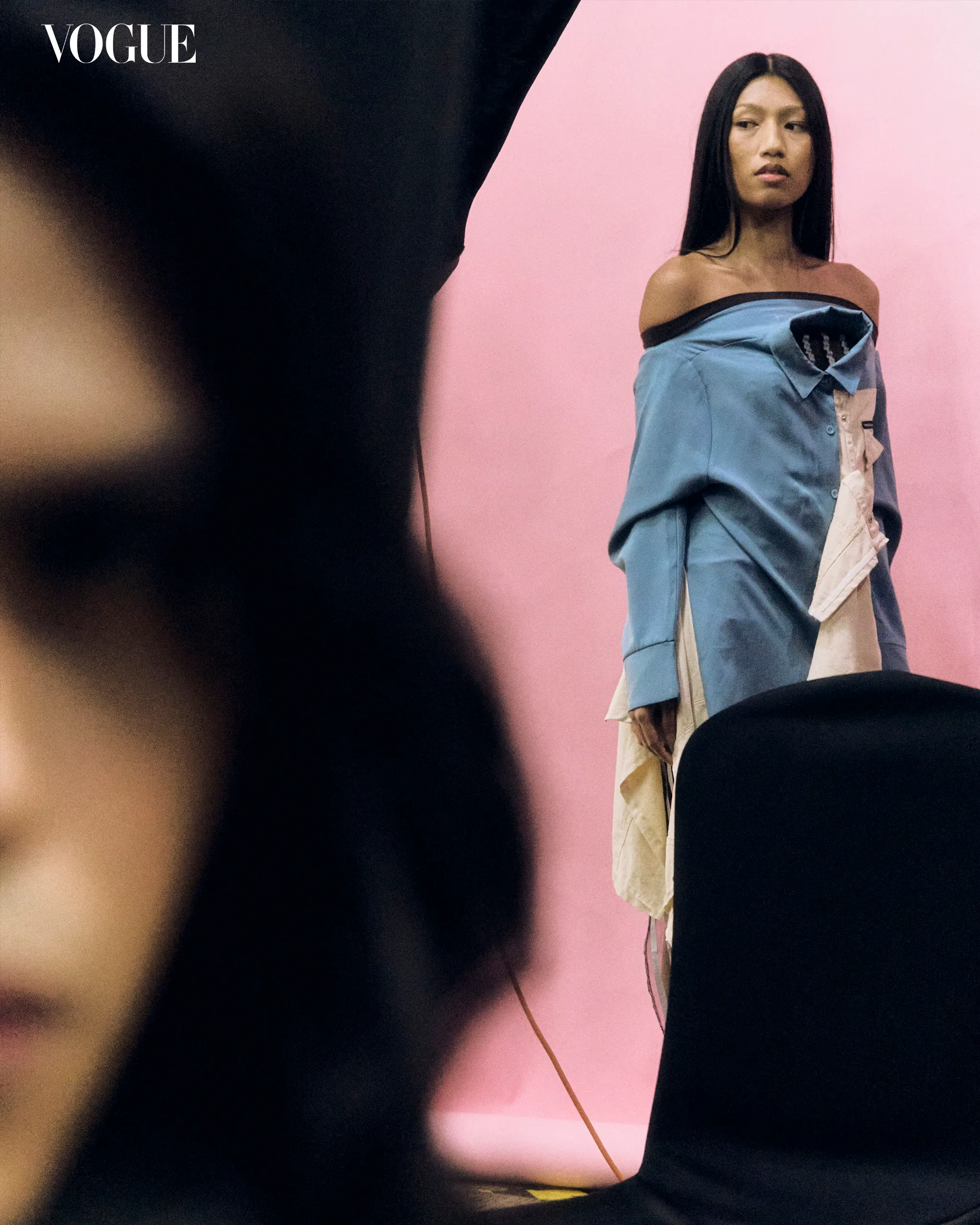Vogue editorial photo: model in off-shoulder blue outfit against pink backdrop, Vogue logo visible in corner, foreground blurred.