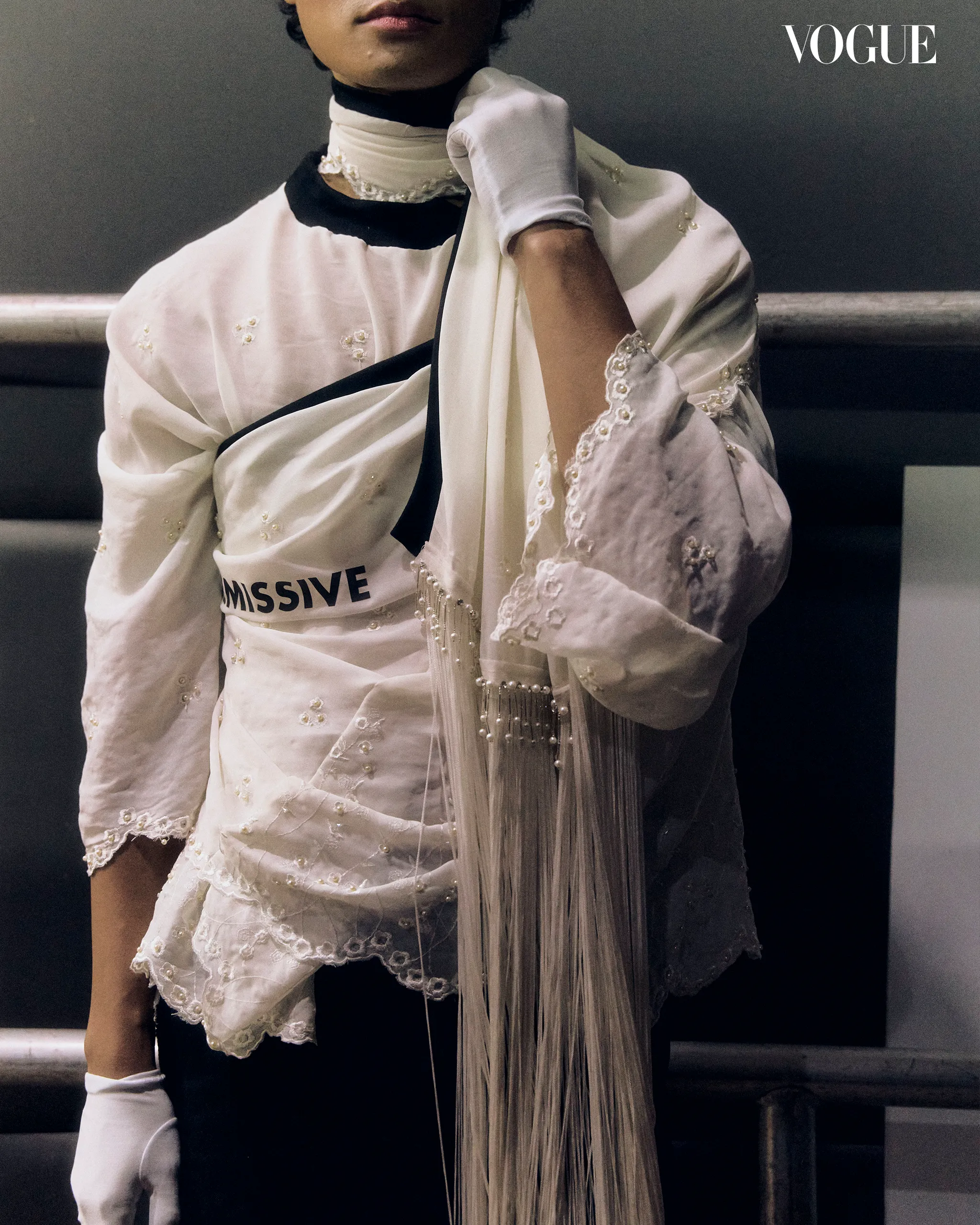Editorial fashion shot: a model in a white embroidered outfit with fringe, wearing a white glove; Vogue logo in the top-right corner.