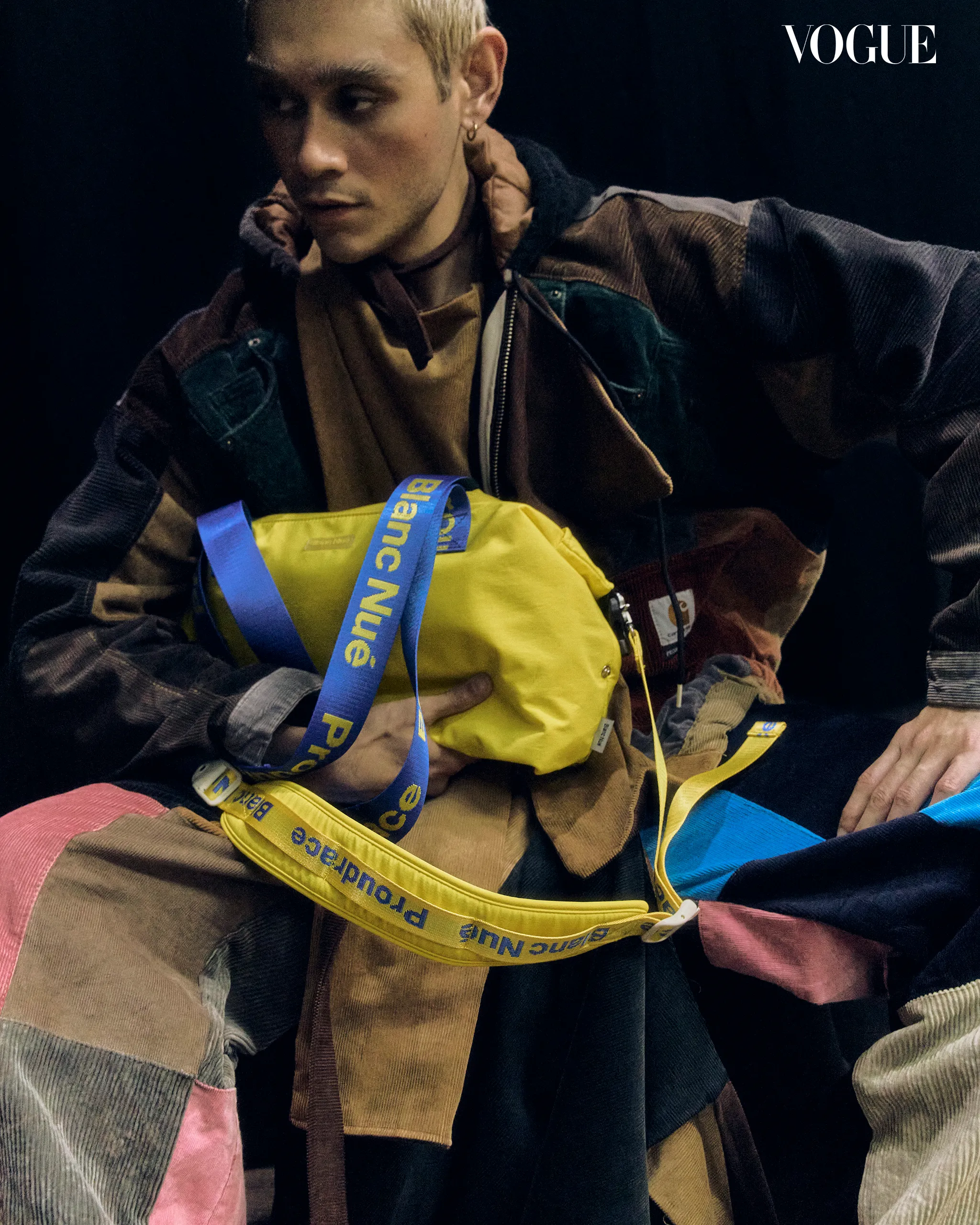 Model wearing a multi‑colored patchwork coat holds a bright yellow bag with blue strap, Vogue magazine logo visible in the top right corner.