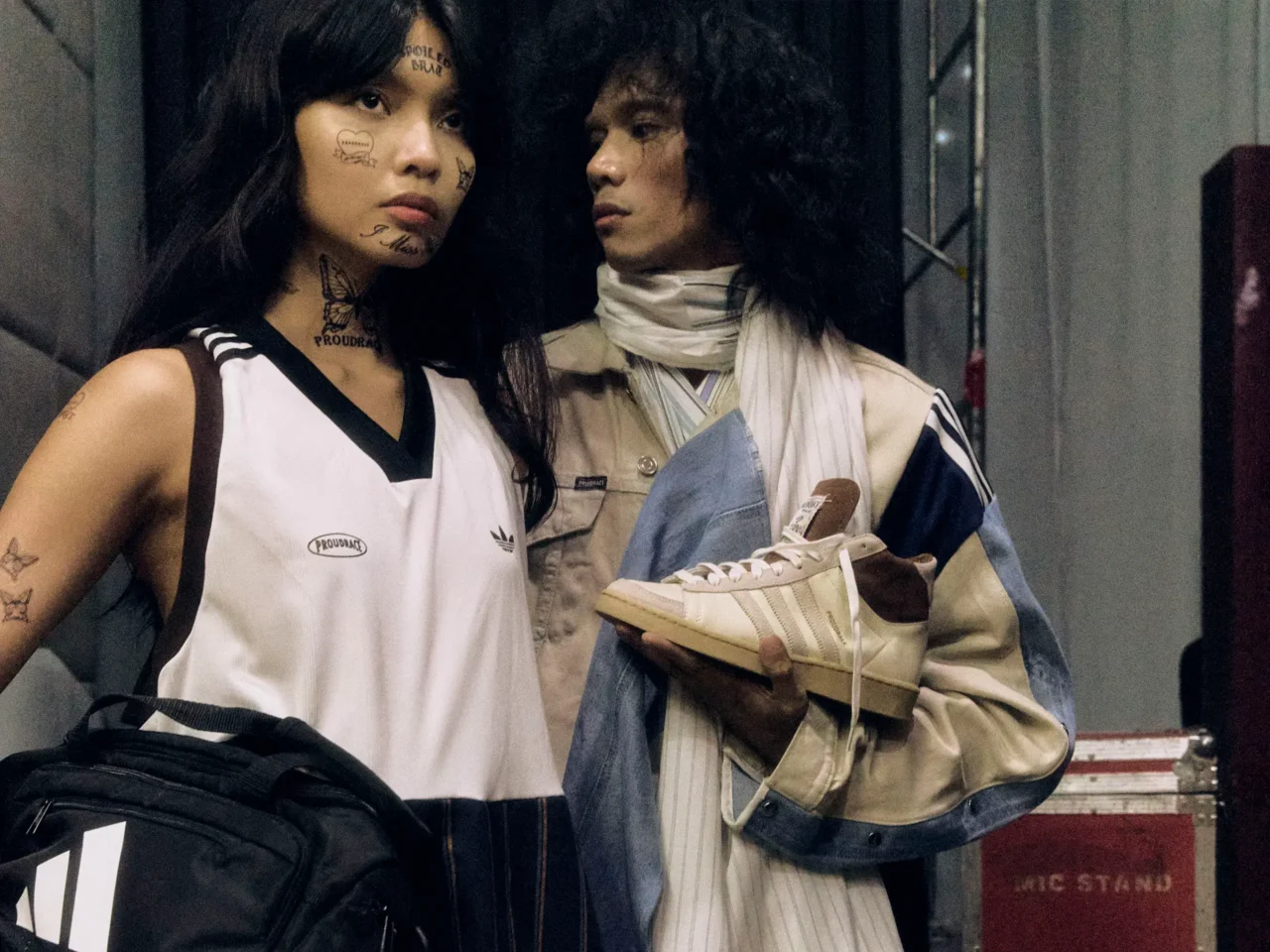 Two women closely posing, one with facial tattoos and a striped outfit, the other in a light jacket and scarf in a dim, urban setting.
