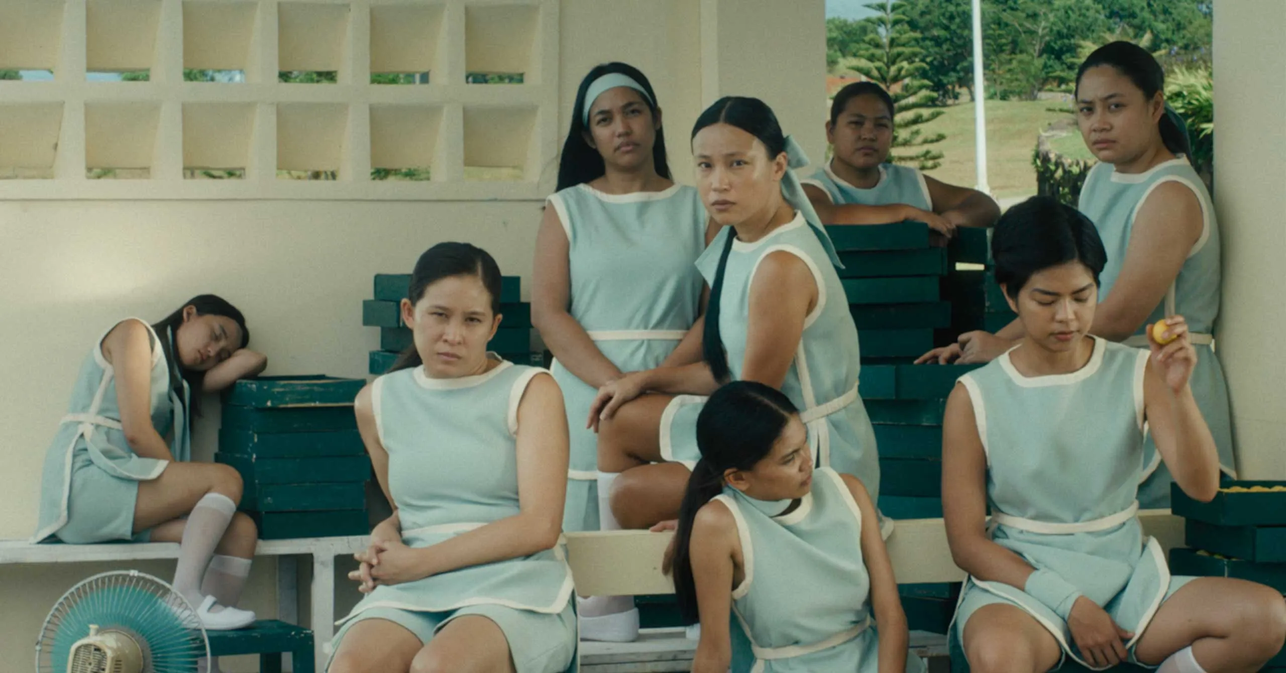 Group of women in matching light blue dresses sit on stacked benches outdoors, some looking toward the camera, a few resting or deep in thought.