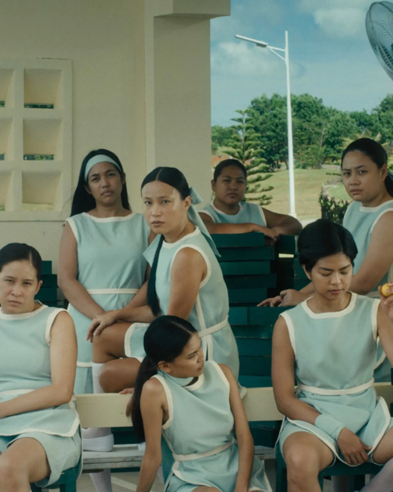 Group of women in matching light blue dresses sit on stacked benches outdoors, some looking toward the camera, a few resting or deep in thought.