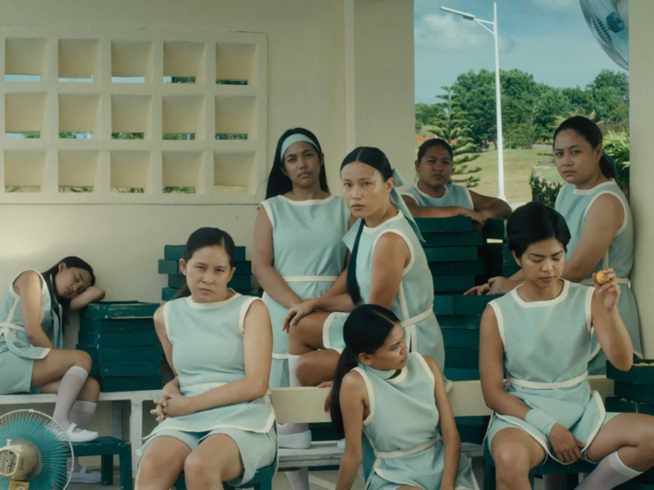 Group of women in matching light blue dresses sit on stacked benches outdoors, some looking toward the camera, a few resting or deep in thought.