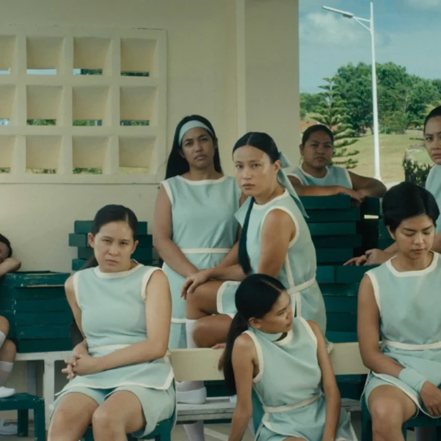 Group of women in light blue uniforms sitting on green bleachers under a shaded area, outdoors.