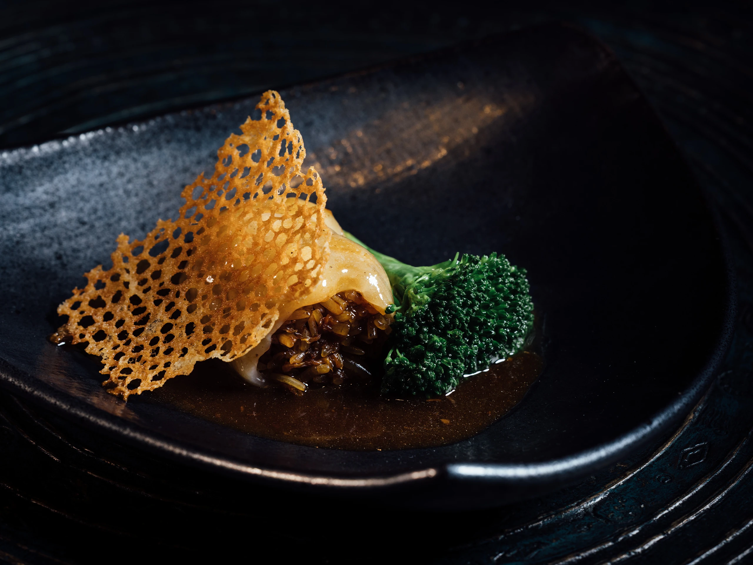 Crispy golden lattice chip placed over a glossy sauce with broccoli florets on a dark plate, close-up view.