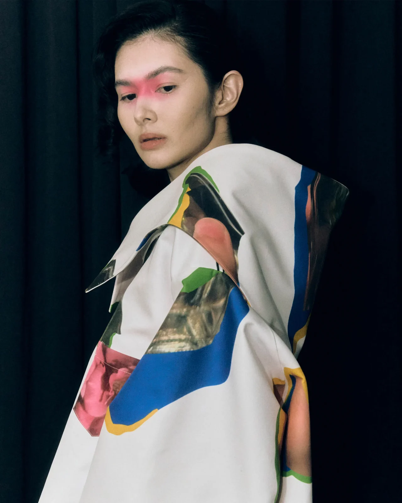 Fashion portrait of a young person with slicked-back dark hair, wearing a black laced shirt with white embroidery, standing against a wooden wall with blue makeup and 'RANDOLFE' written across the forehead.