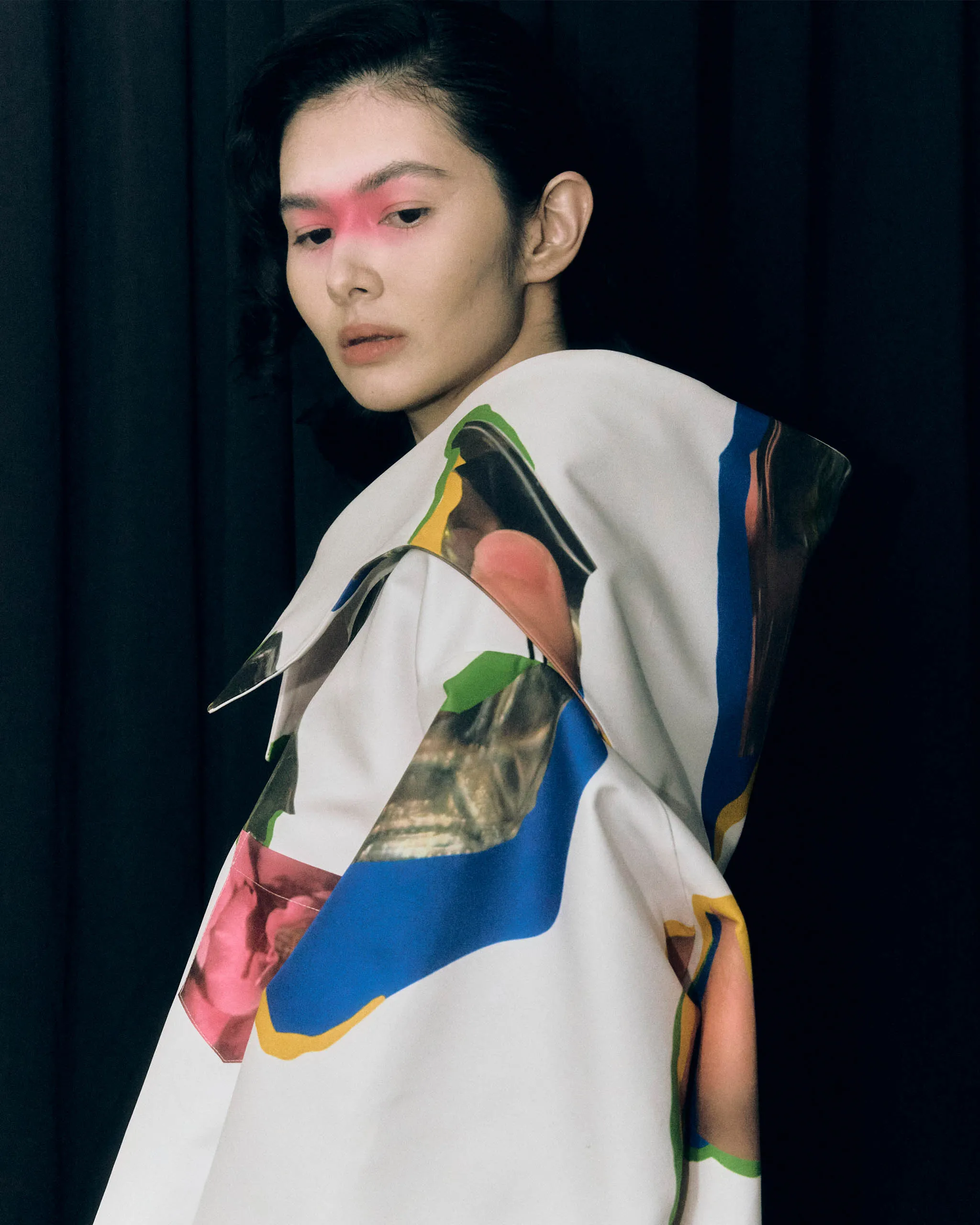 Portrait of a person with bold red eyeshadow spanning the eyelids, looking downward, wrapped in a colorful abstract garment against a dark backdrop.