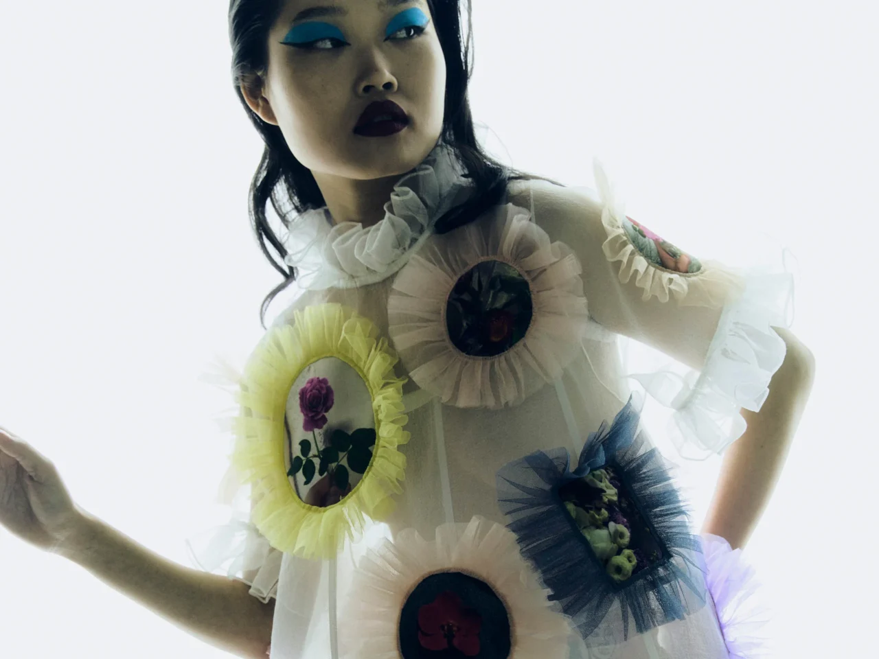Fashion portrait of a young person with slicked-back dark hair, wearing a black laced shirt with white embroidery, standing against a wooden wall with blue makeup and 'RANDOLFE' written across the forehead.