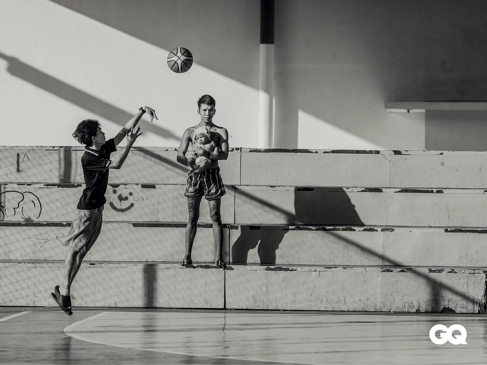 Two young men play basketball on an outdoor court; one leaps to block the ball, the other stands holding a baby.