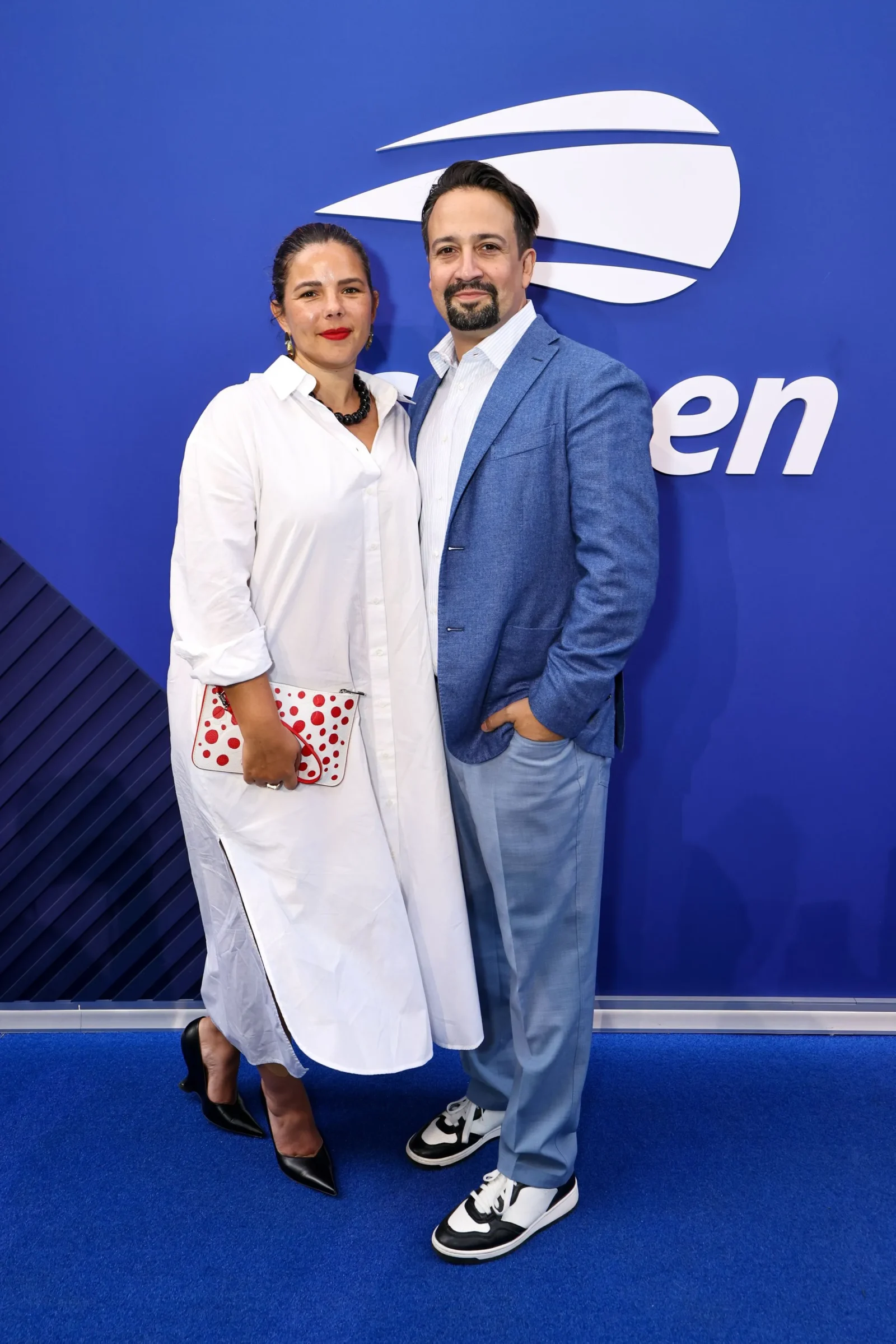 Two adults posing together against a blue wall with a white logo; woman in a white dress holding a red-dotted clutch, man in a blue blazer and light blue pants beside her.